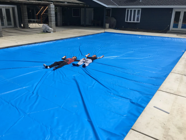 Two people lying on a large blue tarp in a courtyard beside buildings