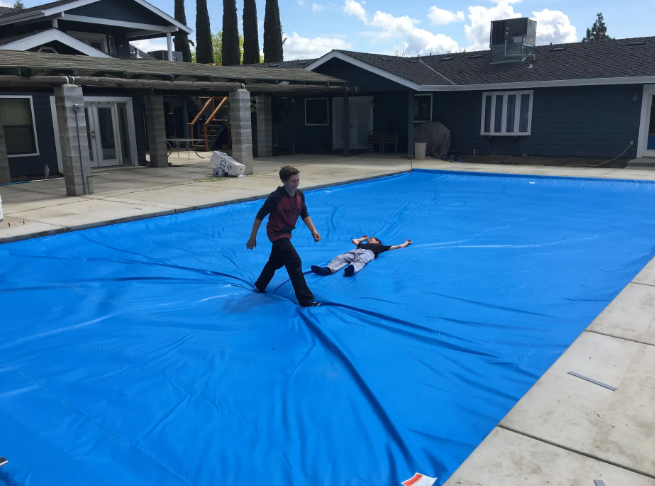Two children on a blue pool cover in a backyard, one walking toward the other near a house.