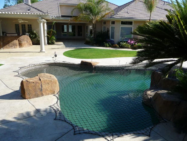 Backyard pool with green net cover, rocks, and a single-story home in the background