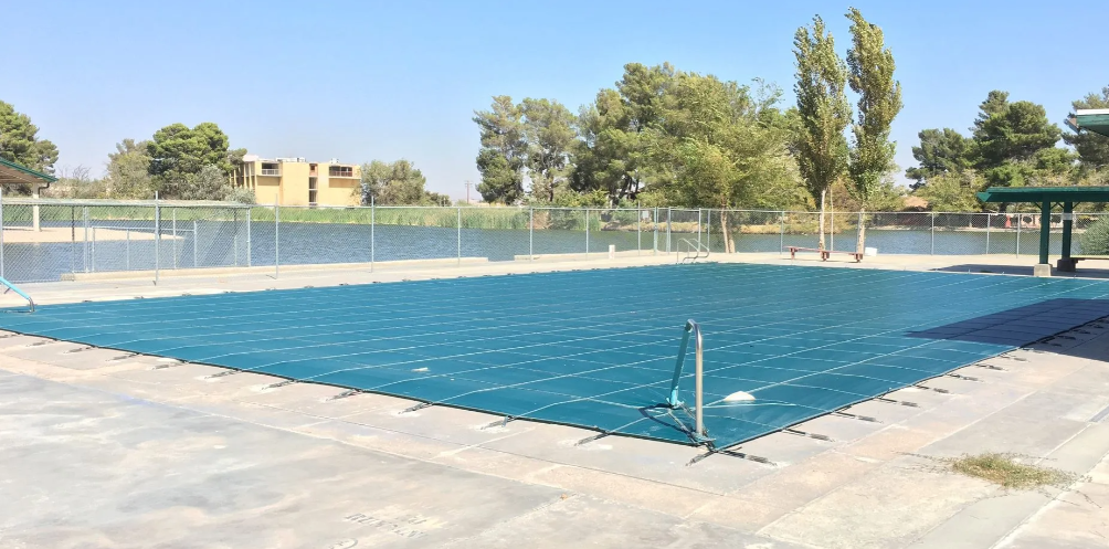 Outdoor swimming pool covered with blue netting, surrounded by a fence and trees under a clear sky
