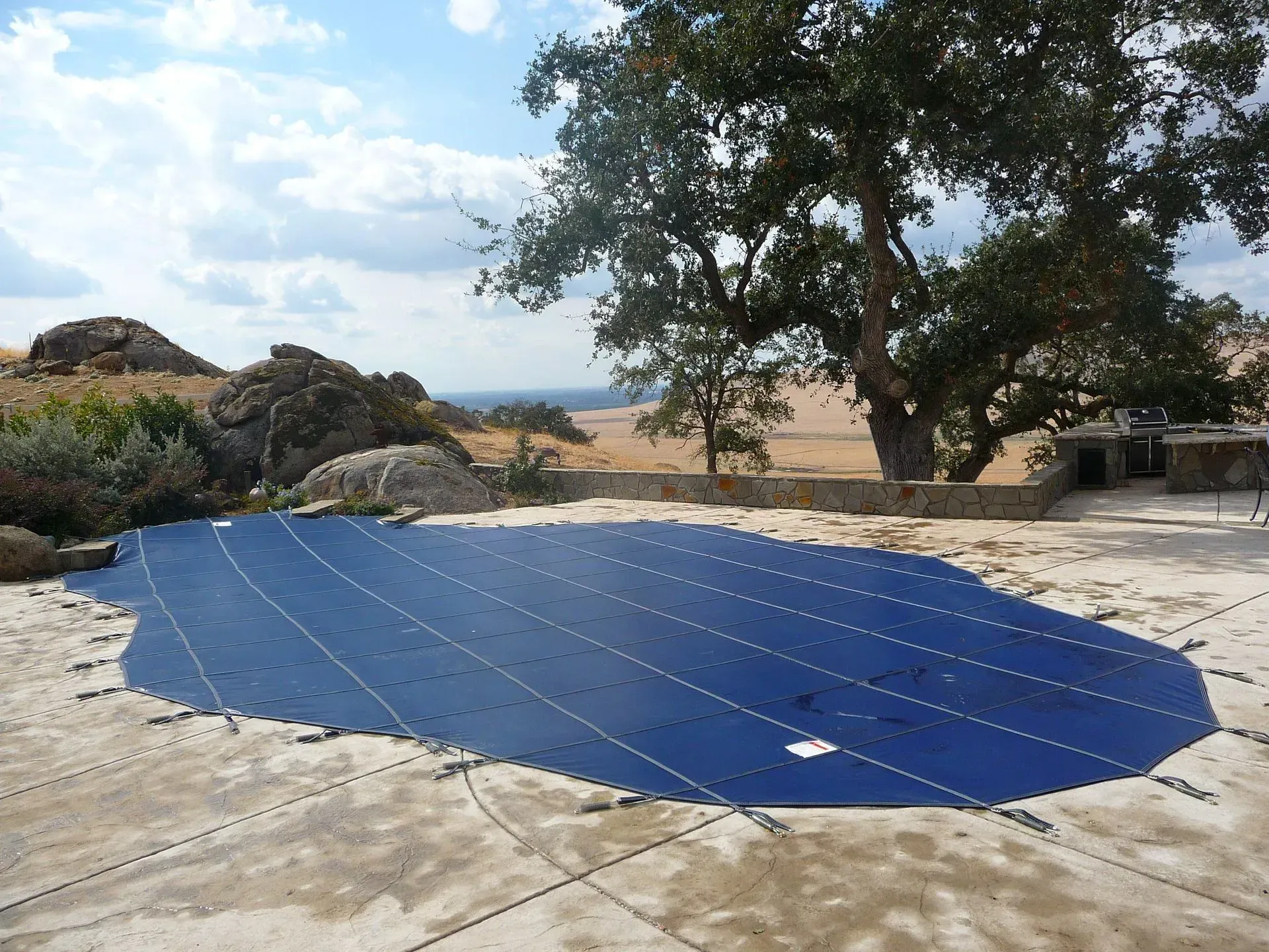 Blue solar panel array on a concrete pad in a rocky outdoor landscape under a tree