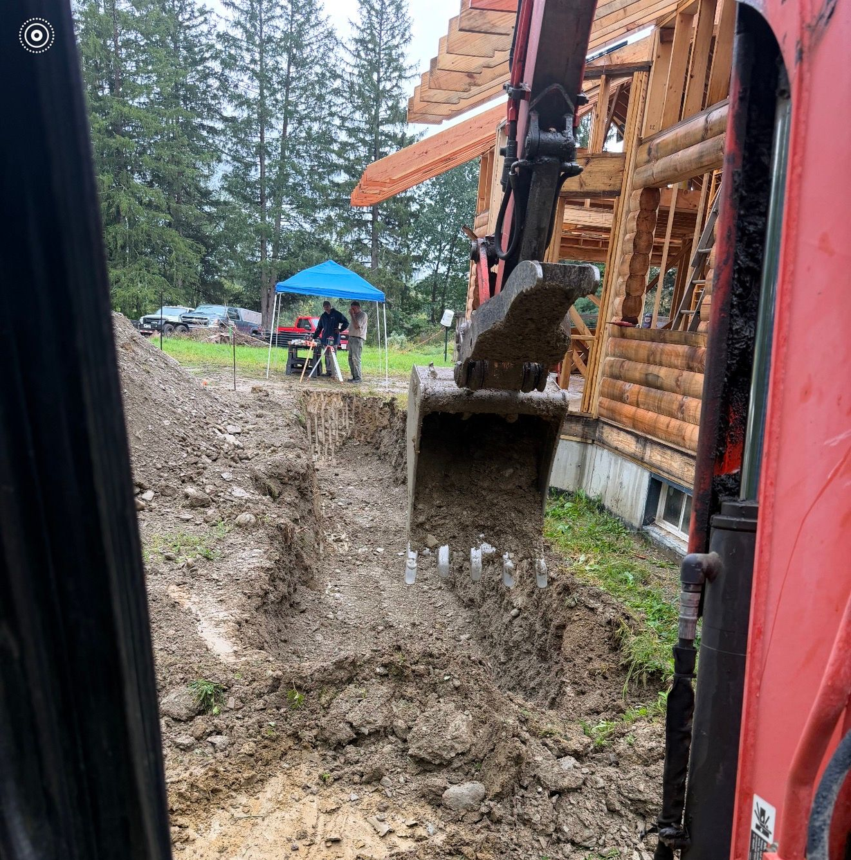 Excavator digging a muddy trench beside a log cabin under construction, with a blue canopy in the background