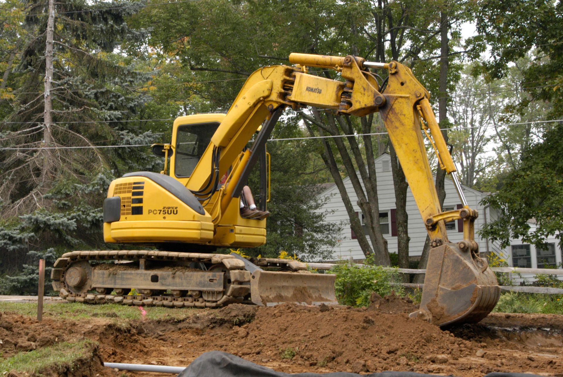 Yellow excavator digging in a dirt area, trees and house in the background.