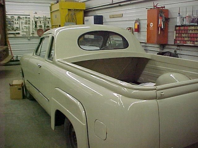 A cream-colored custom car with a pickup truck bed inside a garage. It has an oval rear window.