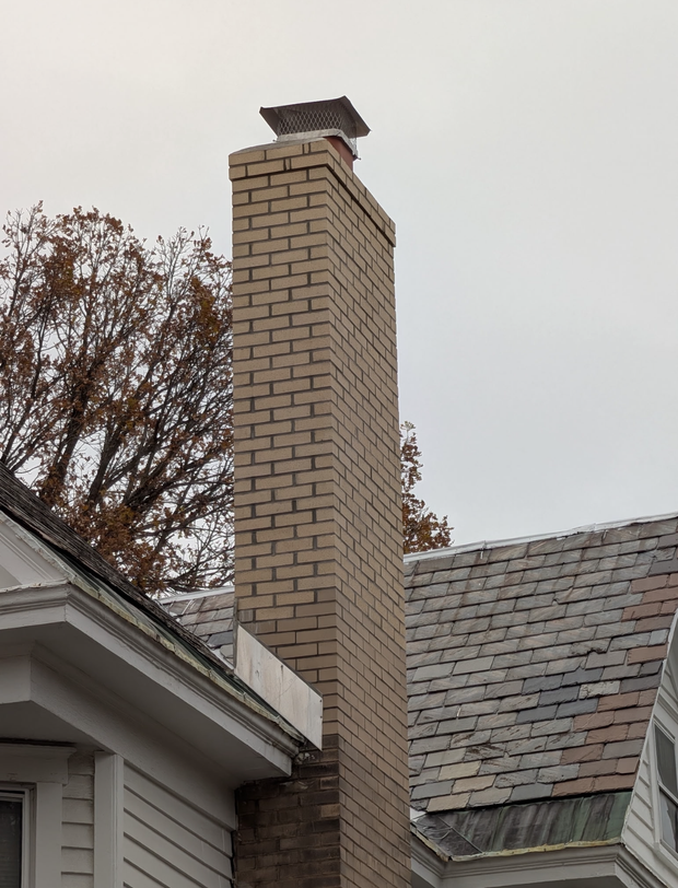 Brick chimney with metal cap on a gray house roof under an overcast sky
