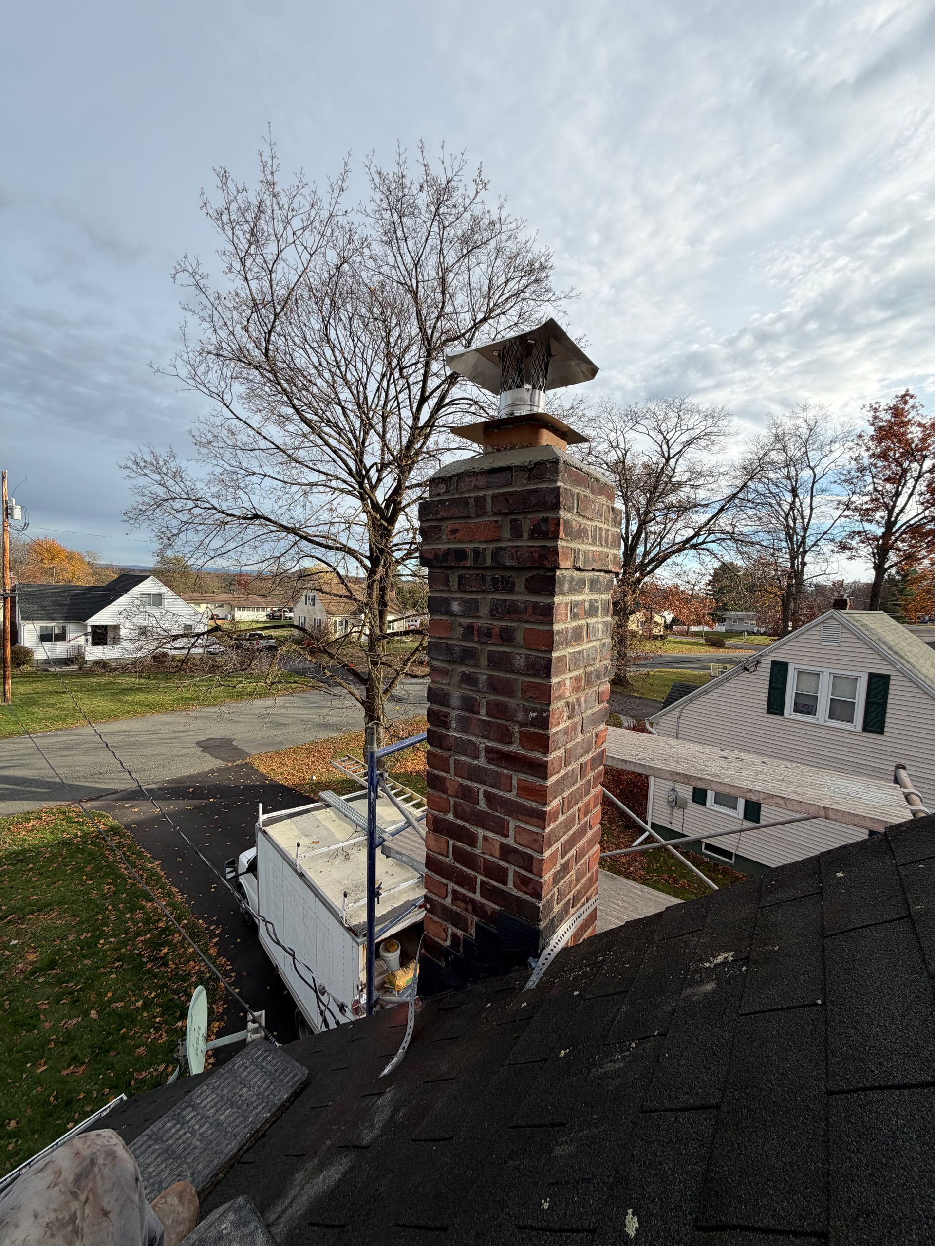 Brick chimney with metal cap on a rooftop, overlooking houses and trees under a cloudy sky