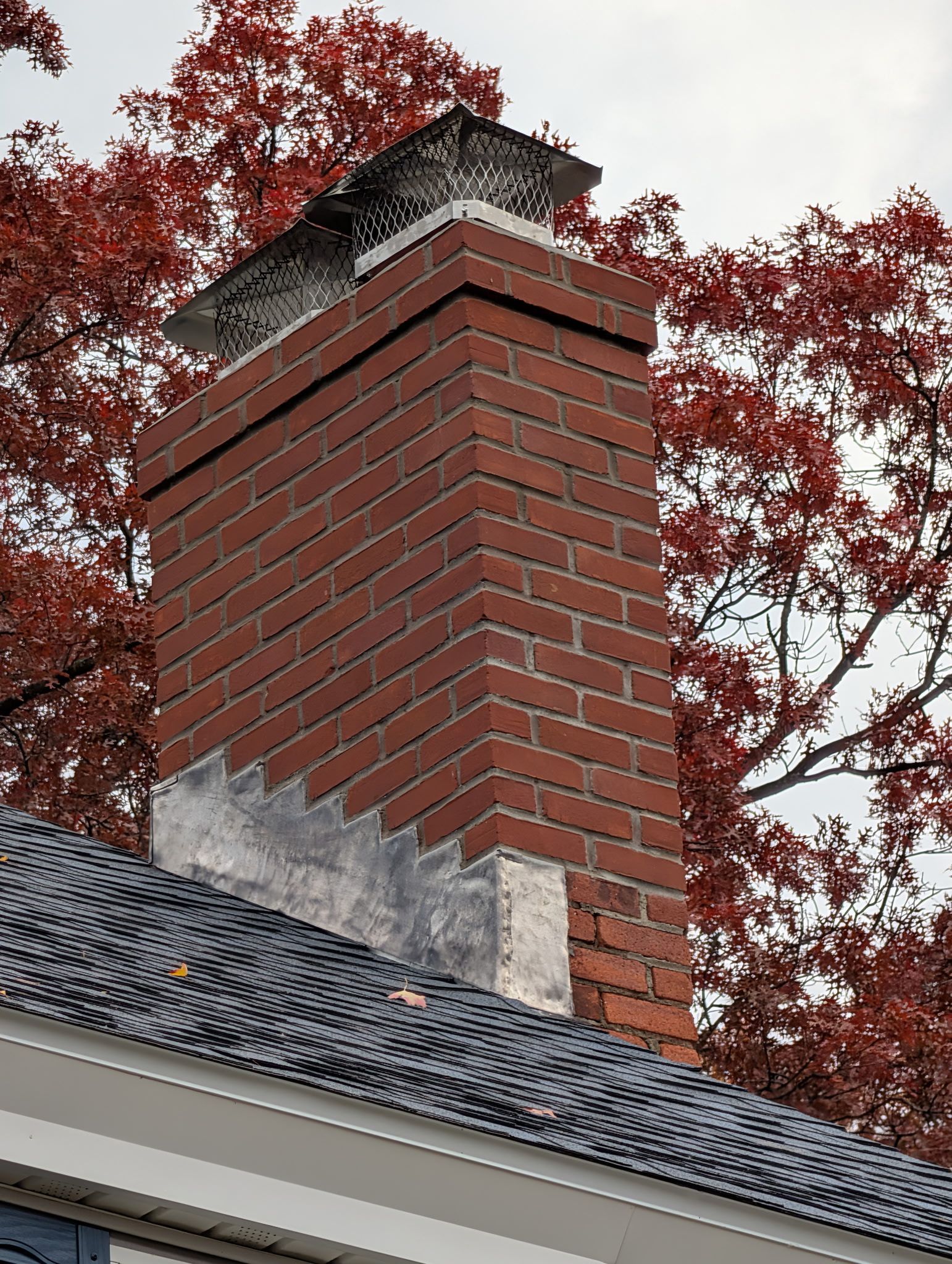 Red brick chimney on a shingled roof with red autumn leaves in the background