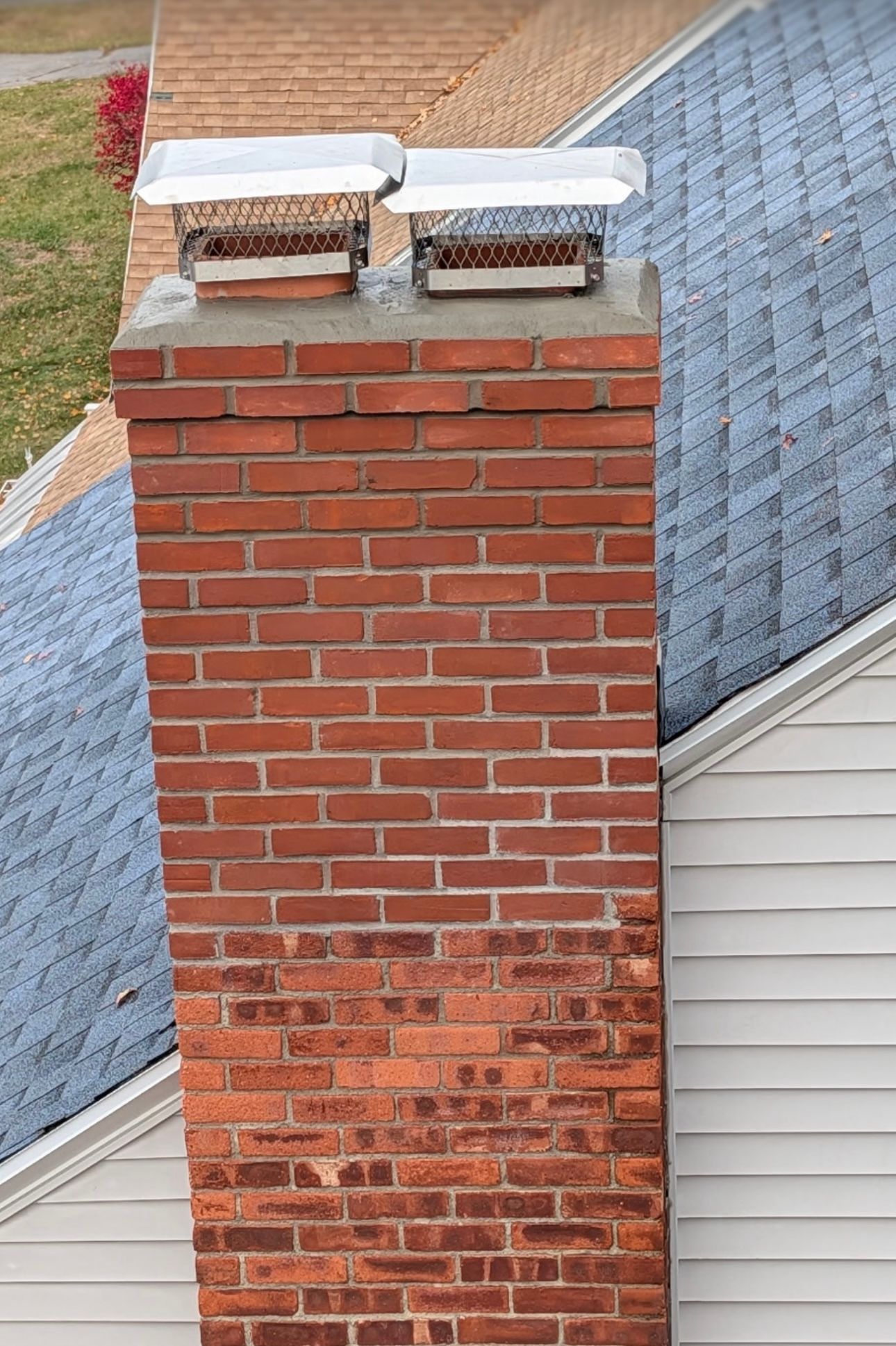 Red brick chimney with two capped vents beside a gray shingled roof and white siding