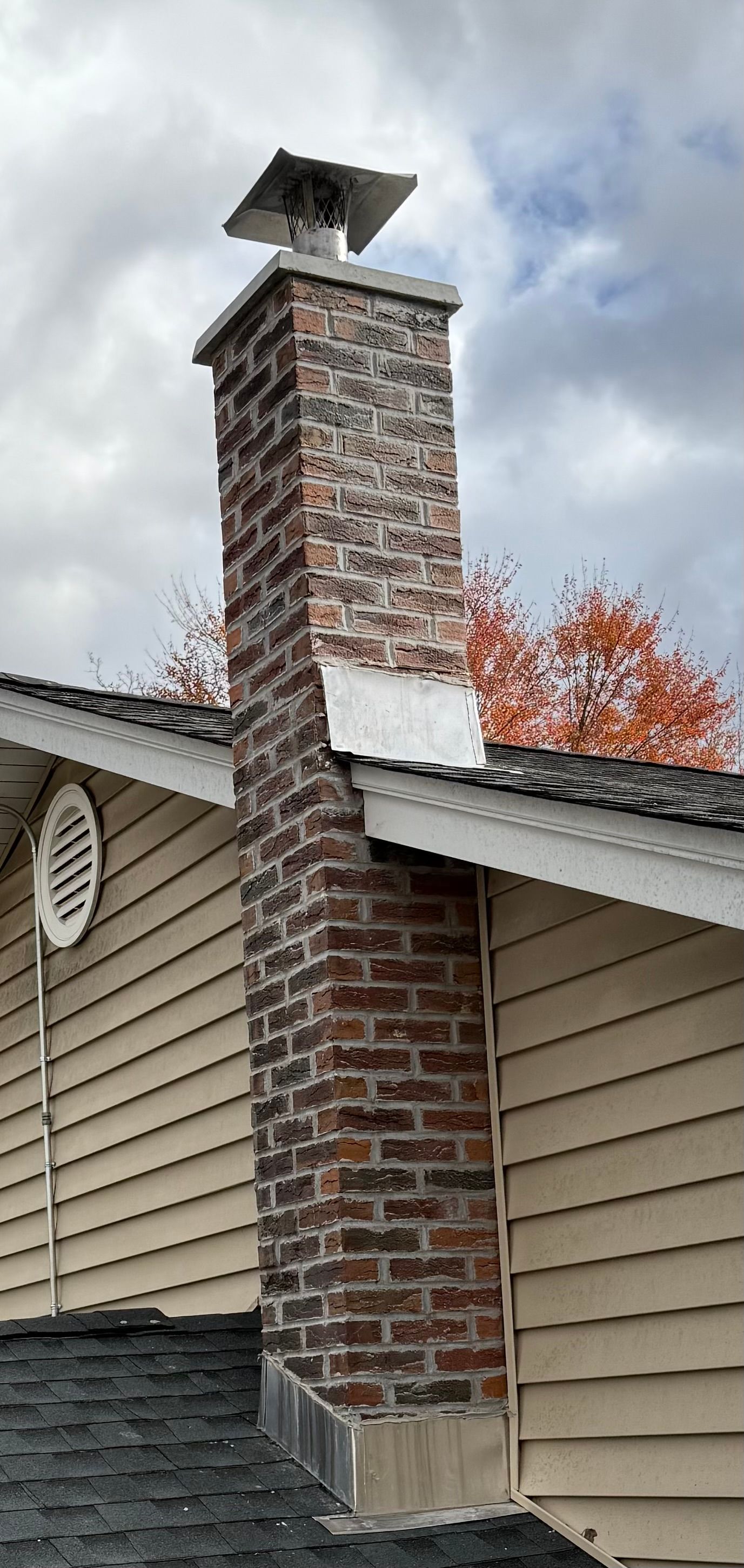 Brick chimney with white cap on a beige house roof against cloudy sky and autumn trees