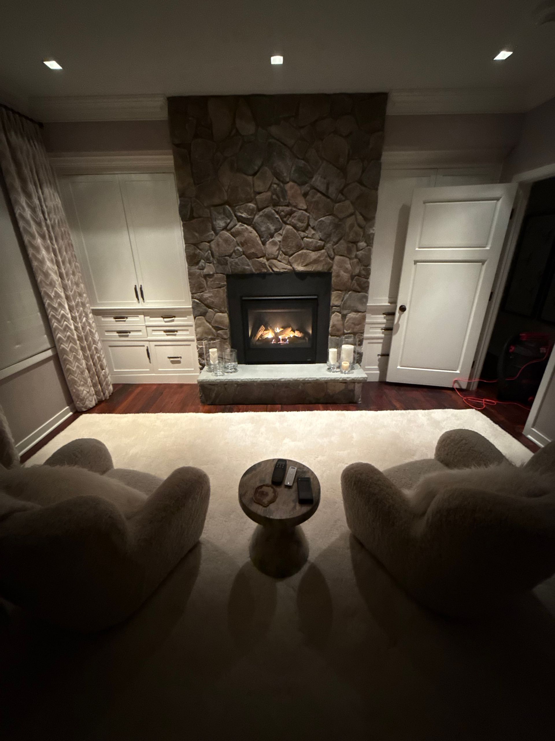 Cozy living room with stone fireplace, white cabinets, two chairs, and a small round table facing the fire.