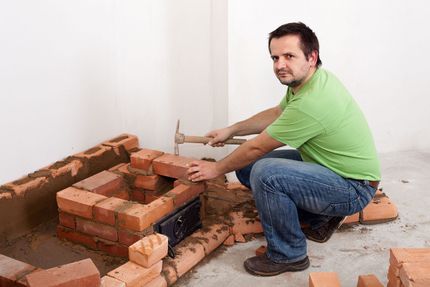 Man kneeling beside a brick structure, using a hammer in a room with scattered bricks and mortar scraps
