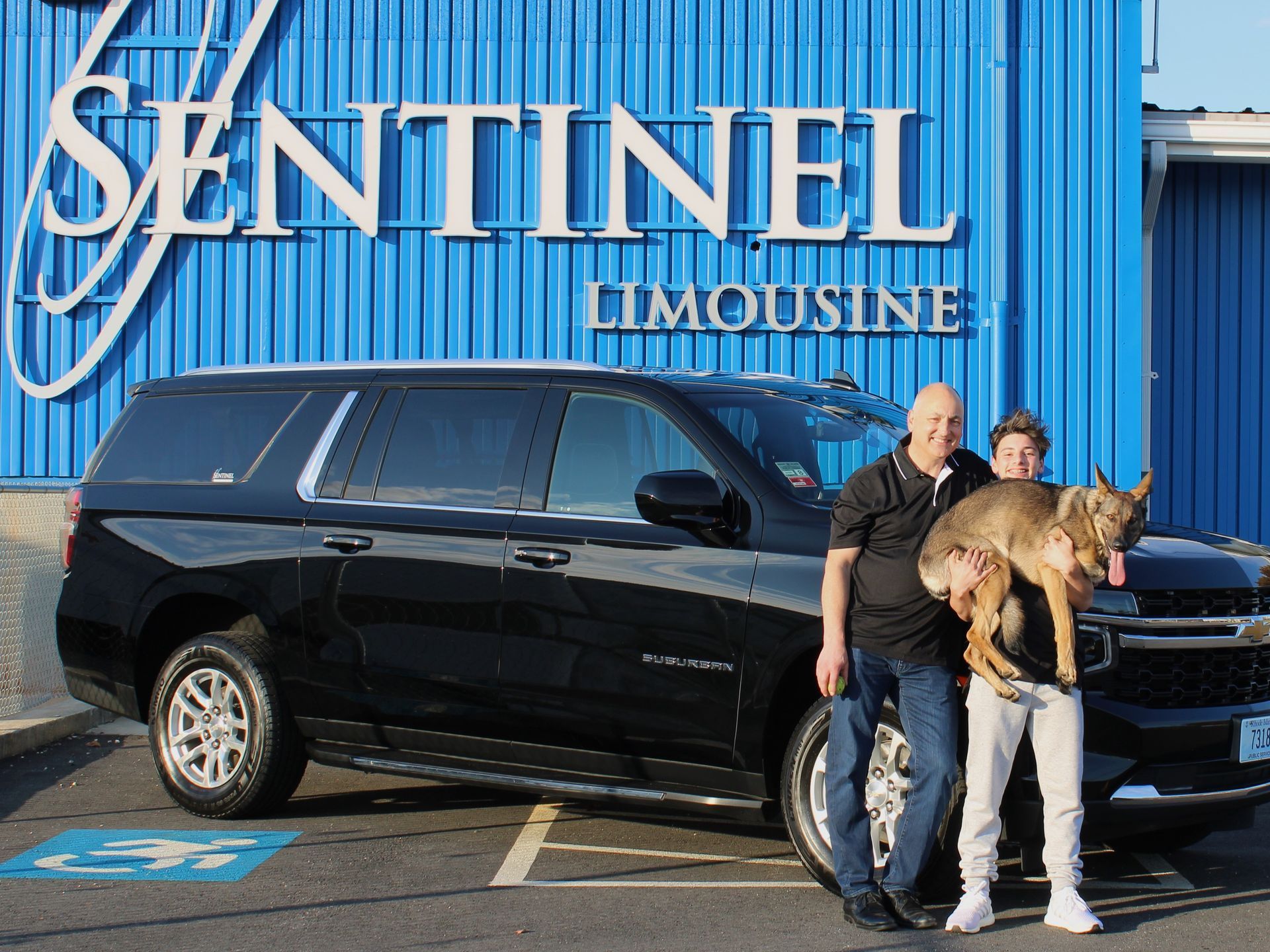 Two men with a dog stand next to a black SUV in front of a blue 