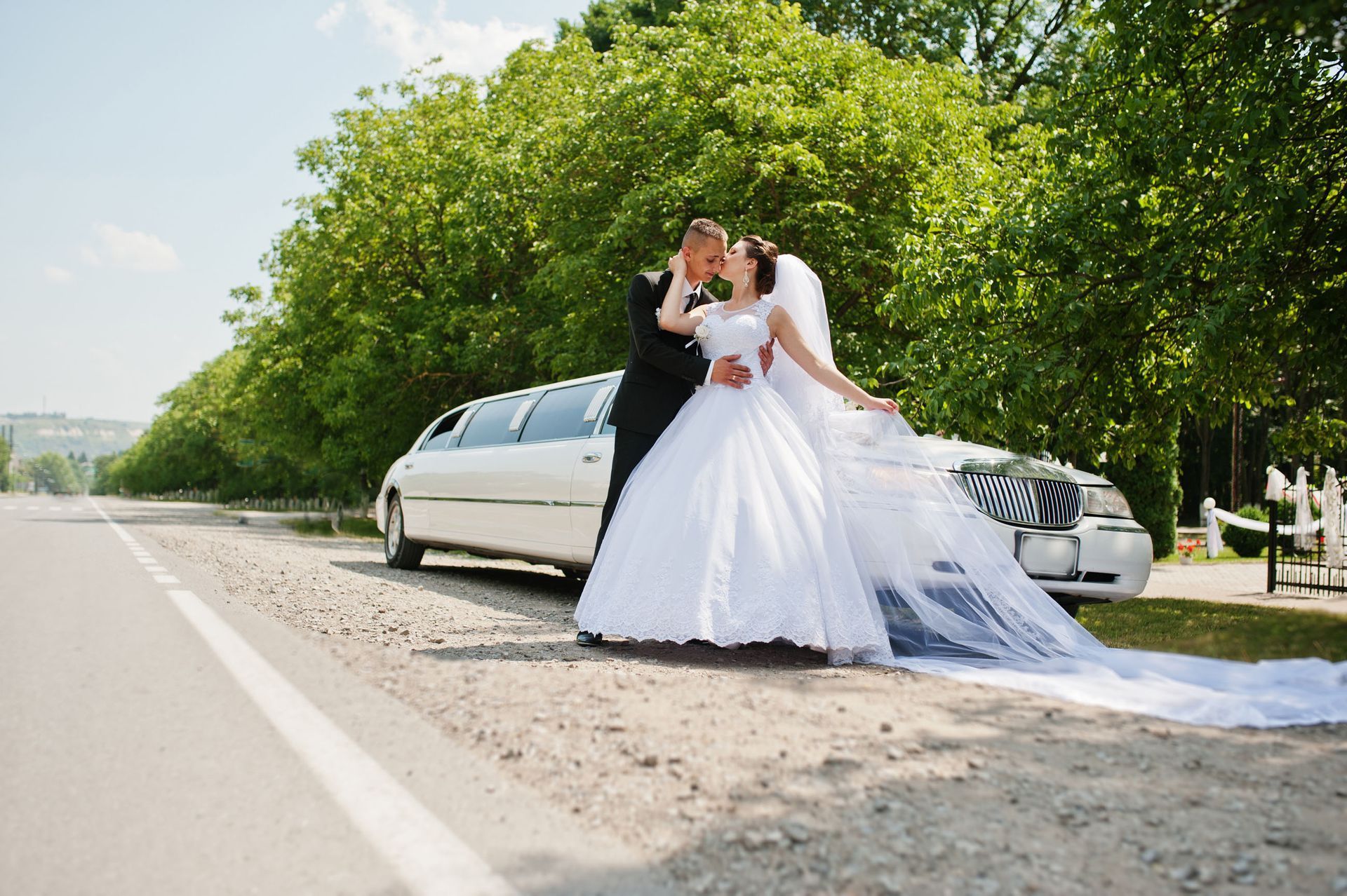 Bride and groom embrace next to a white limousine on a sunny roadside; bride in white gown, groom in black suit.