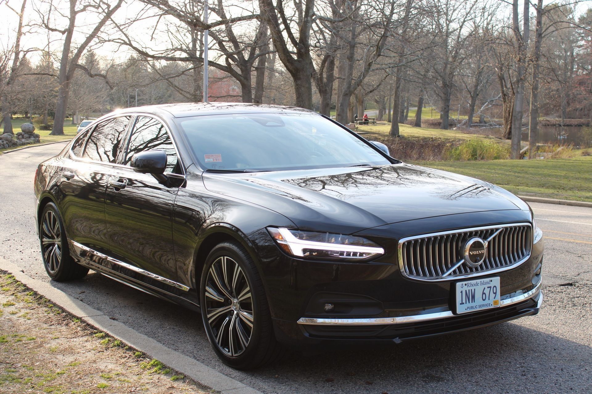 Black Volvo S90 parked on a road next to a grassy area with bare trees.