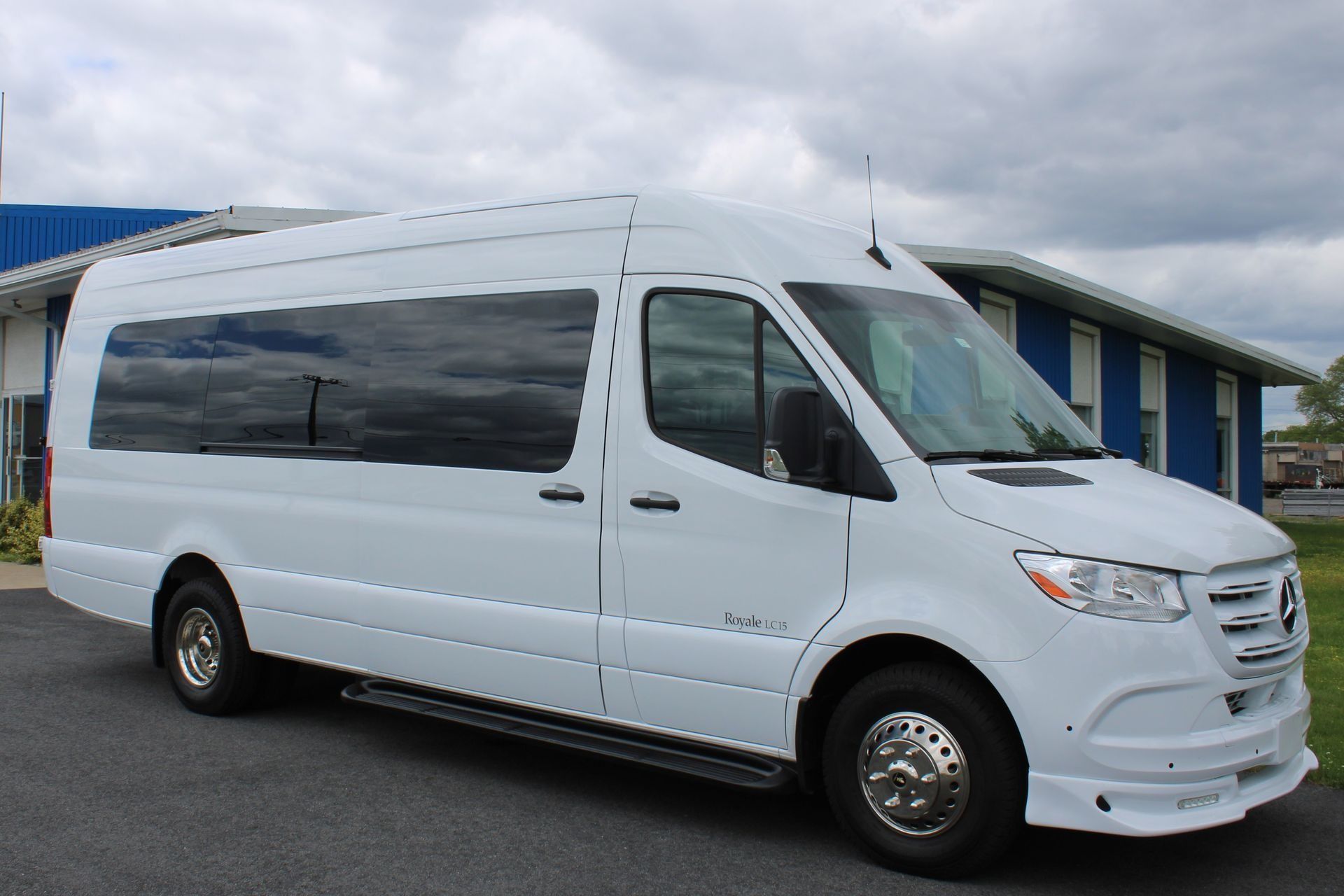 White Mercedes-Benz passenger van parked outside a building with blue and white trim on a cloudy day.