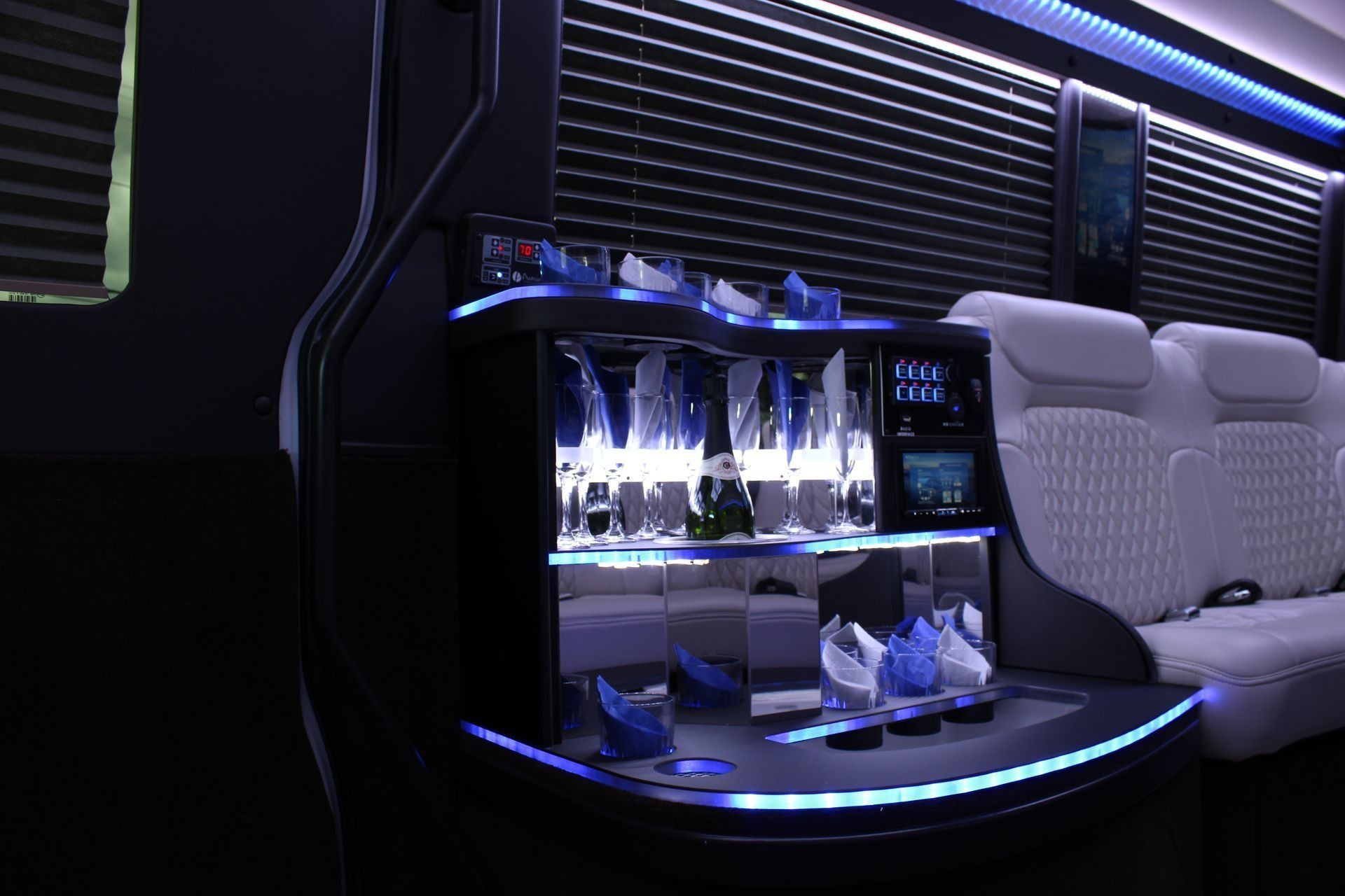 Luxury van interior with illuminated bar, glassware, and white seating; dark walls and blue lighting.