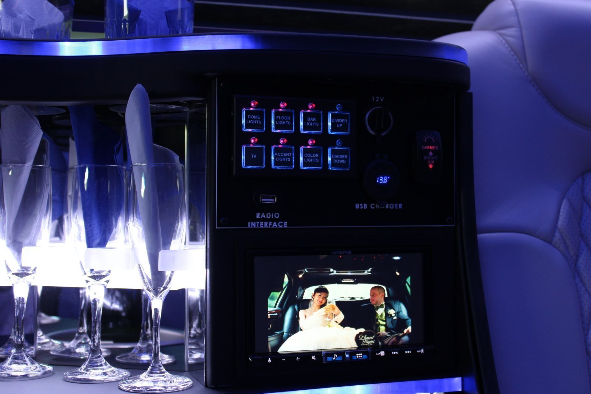 Interior of a limousine, with control panel, champagne flutes, and a screen displaying a wedding couple.
