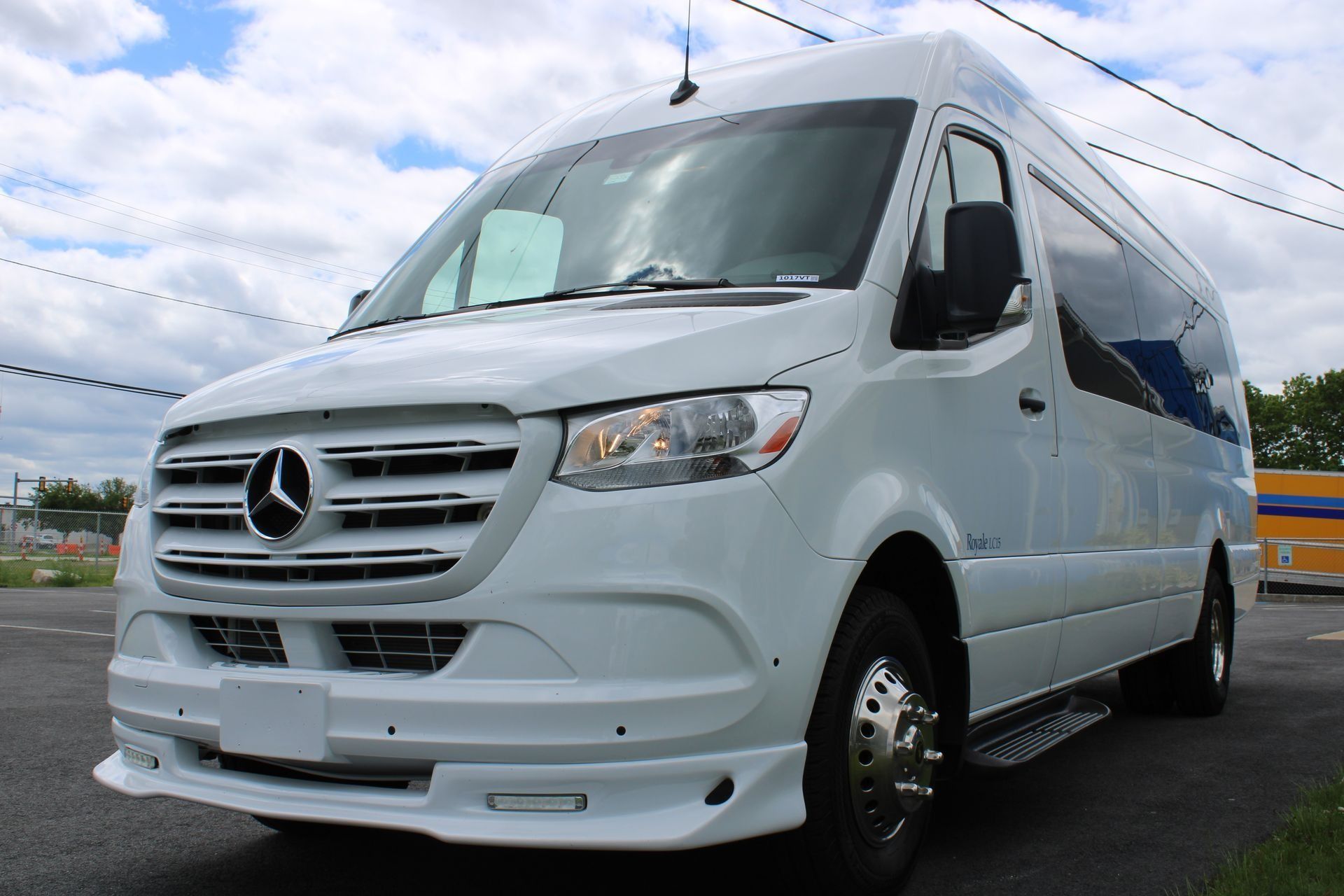 White Mercedes Sprinter passenger van parked on a paved road, partly cloudy sky.