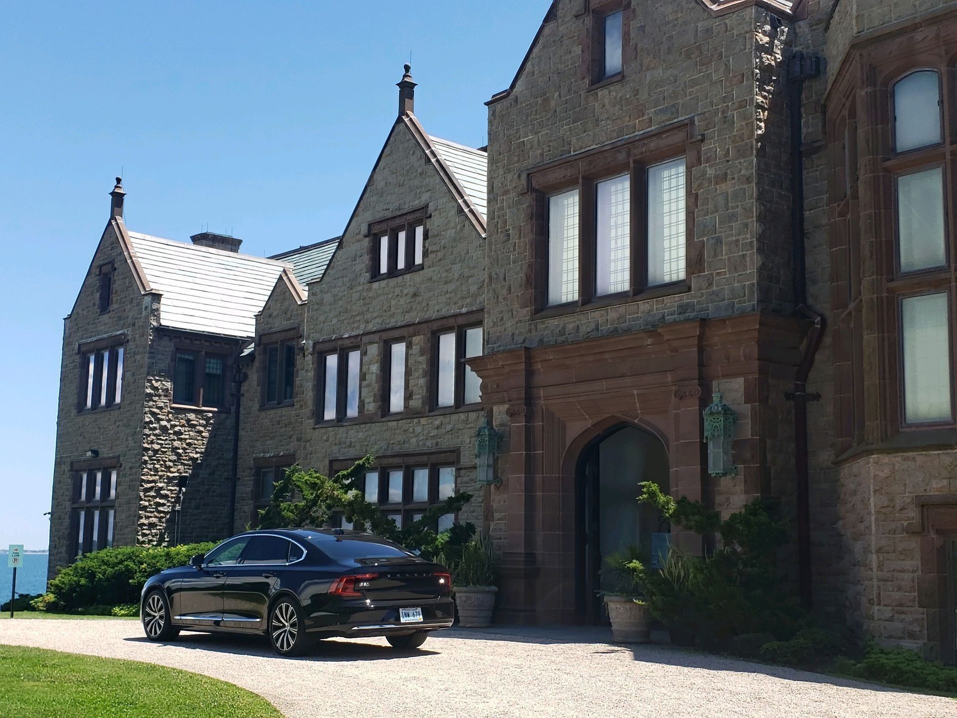 Black car parked in front of a large stone mansion with a blue sky background.