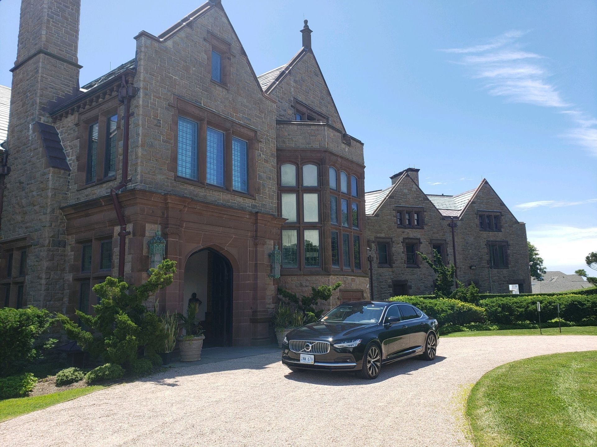 A dark car parked in front of a large stone mansion with a gravel driveway on a sunny day.