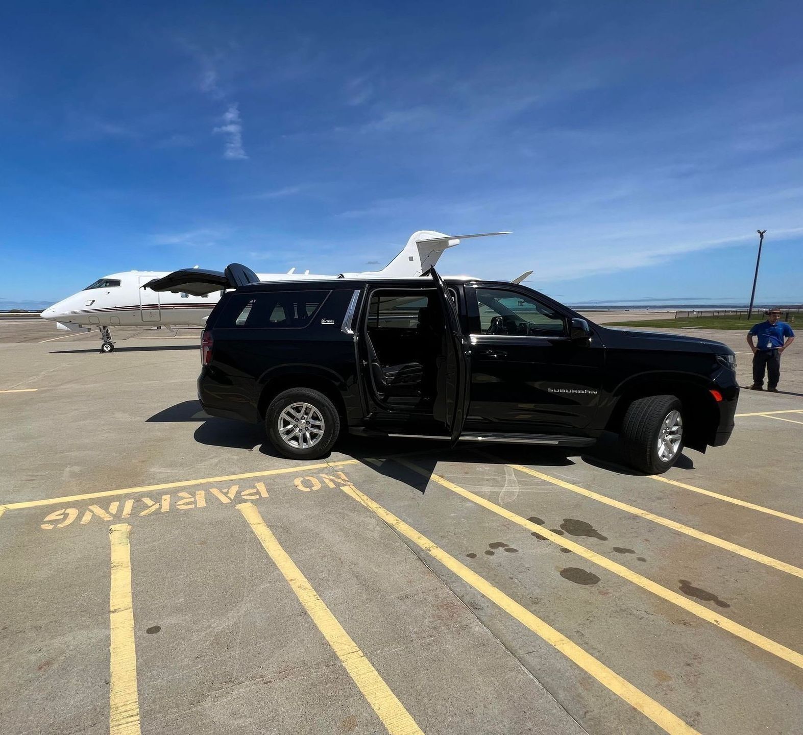 Black SUV with open doors near a private jet on a tarmac under a blue sky.