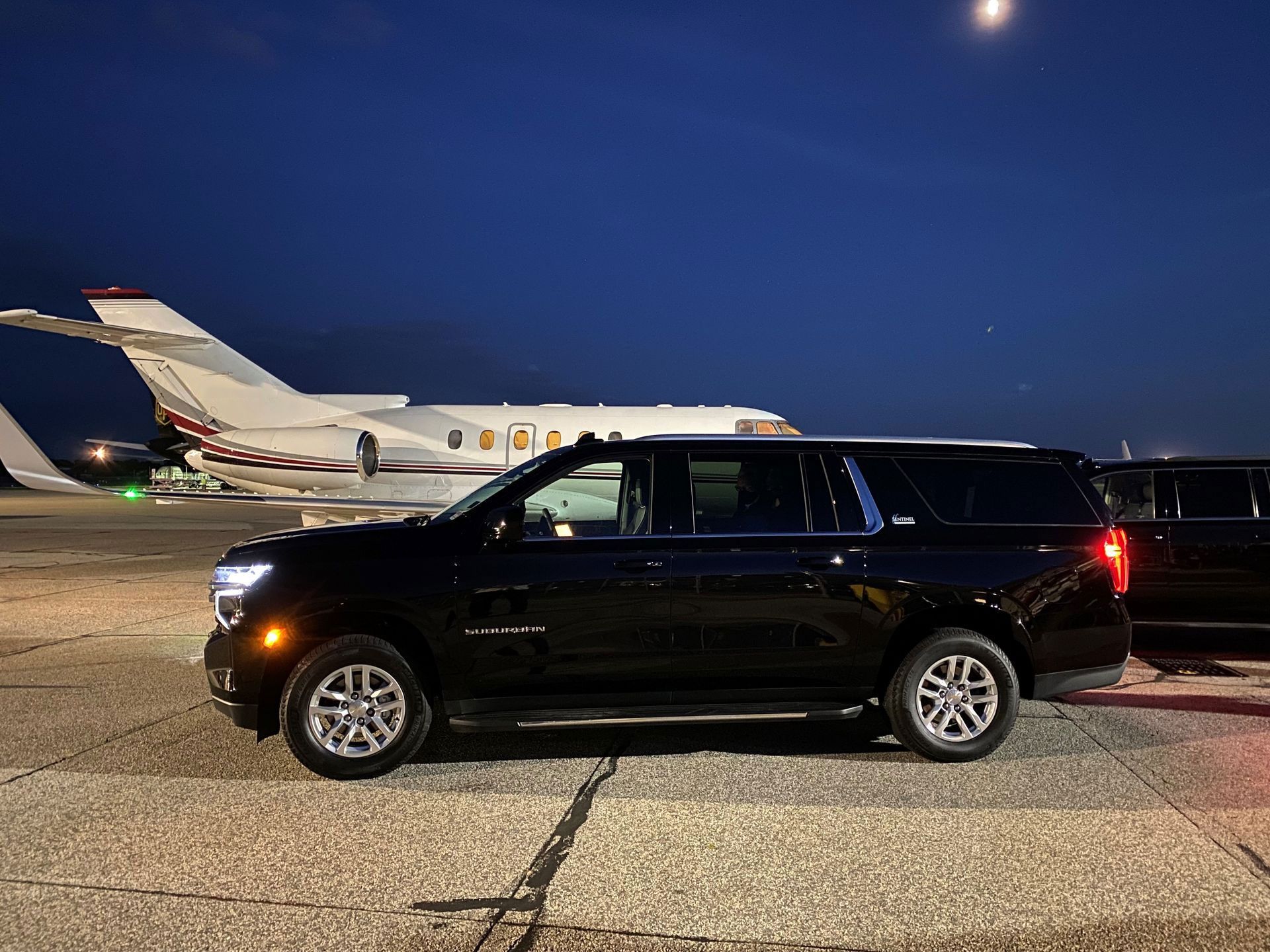Black SUV on an airport tarmac with a private jet in the background at night.