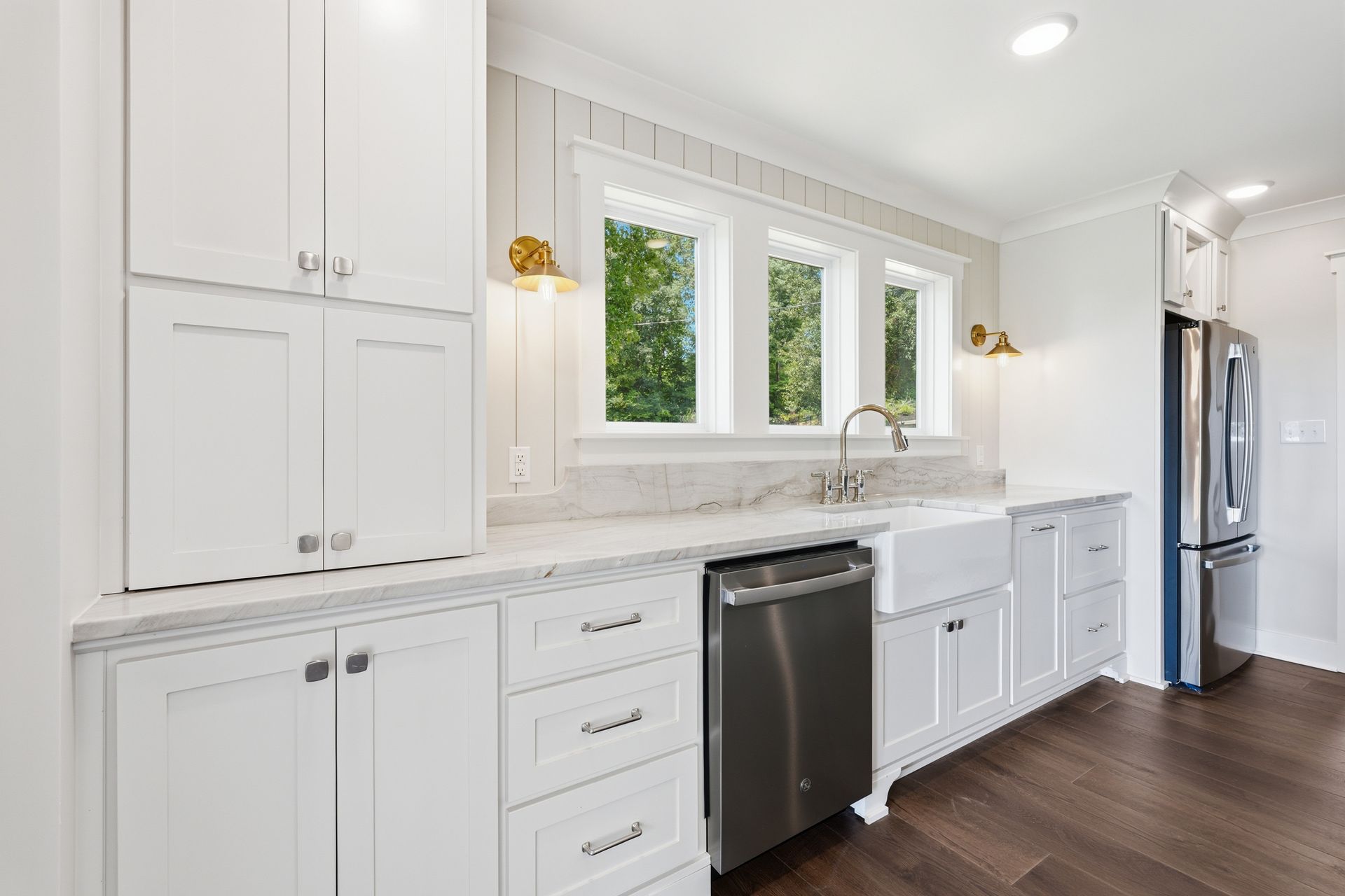 Bright white kitchen with marble counters, black dishwasher, and stainless steel fridge under three windows