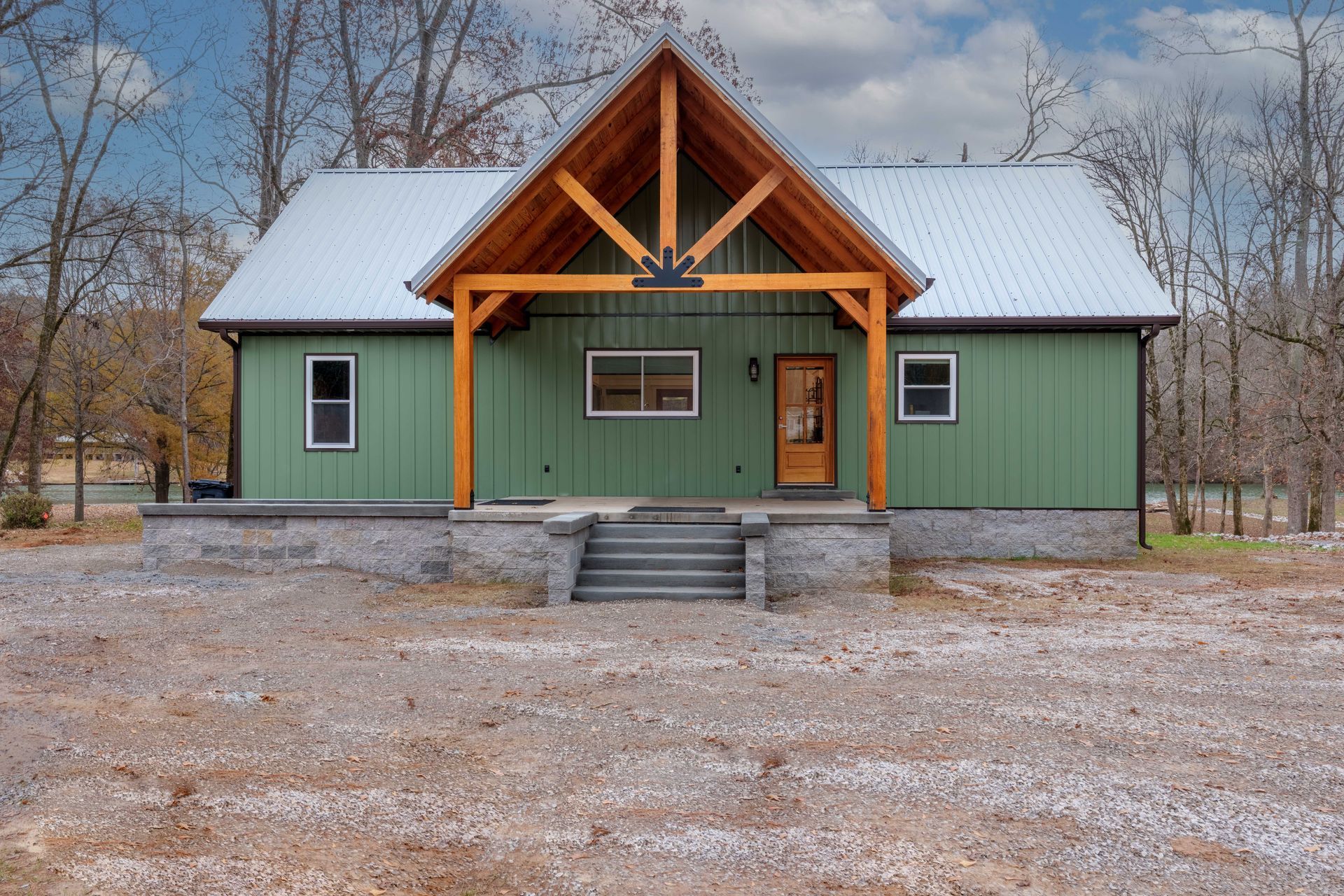 Green cabin with a snow-dusted roof and orange porch trim, centered in a wooded winter setting