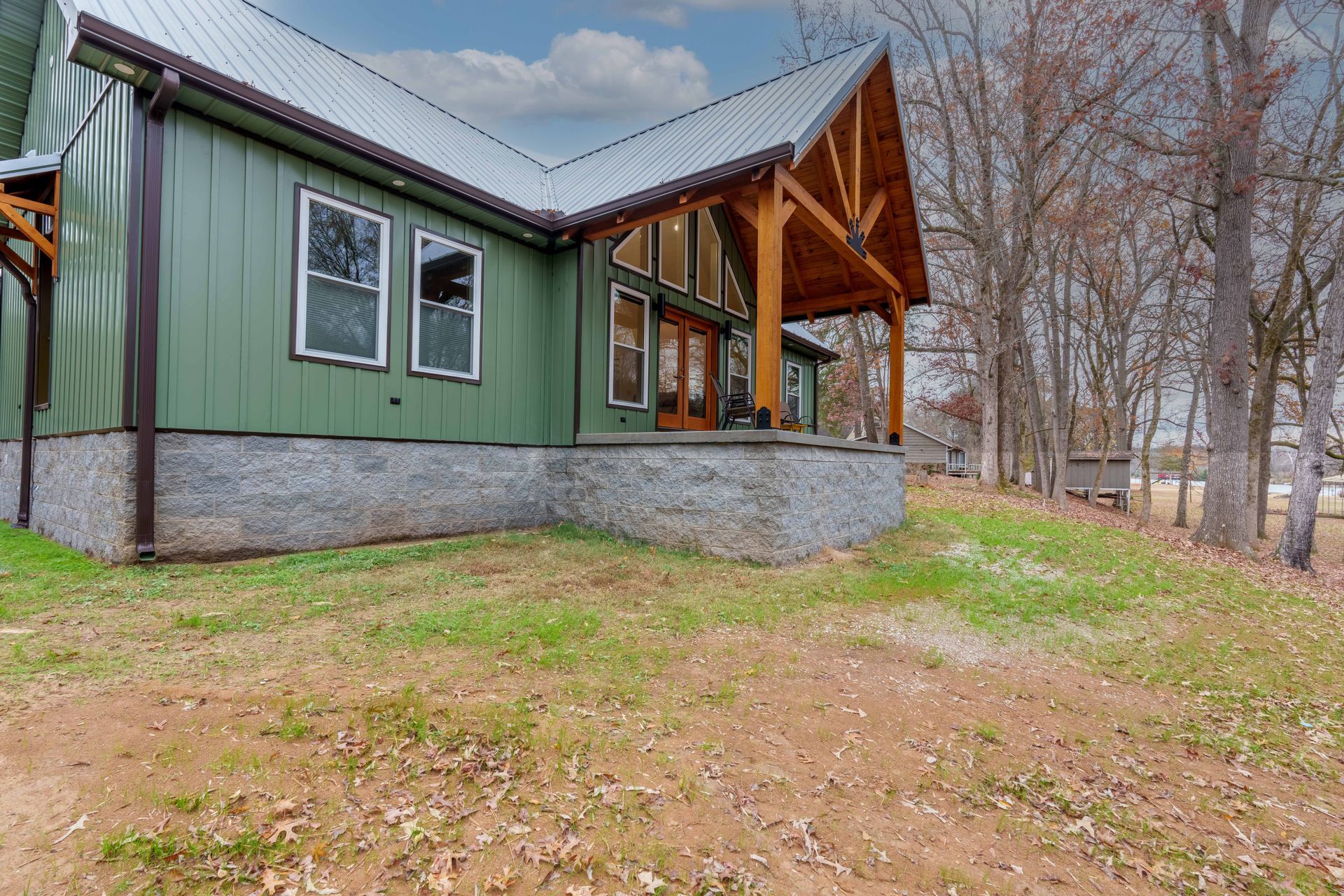 Green cabin with stone foundation and wooden porch in a wooded yard