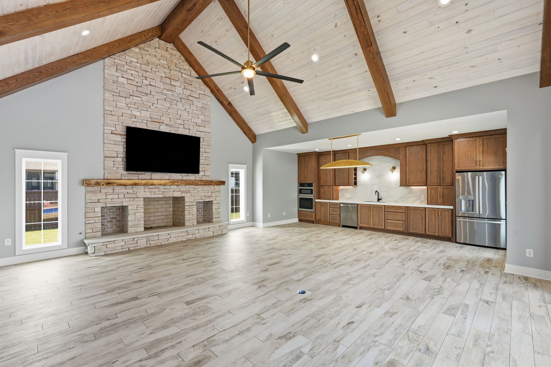 Bright open living room with vaulted ceiling, stone fireplace, large TV, and an open kitchen with wood cabinets.