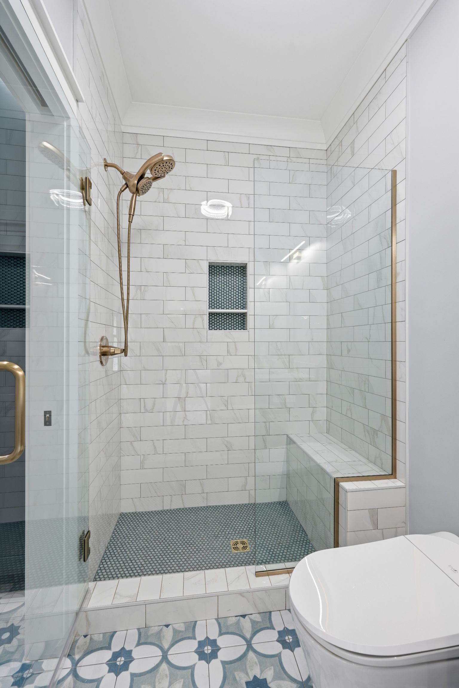 Compact white-tiled bathroom with glass shower, built-in bench, and patterned floor tile.