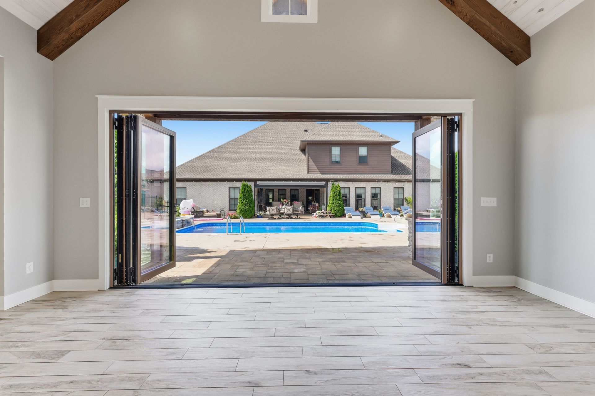 Open room with vaulted ceiling facing a pool and house through wide glass doors