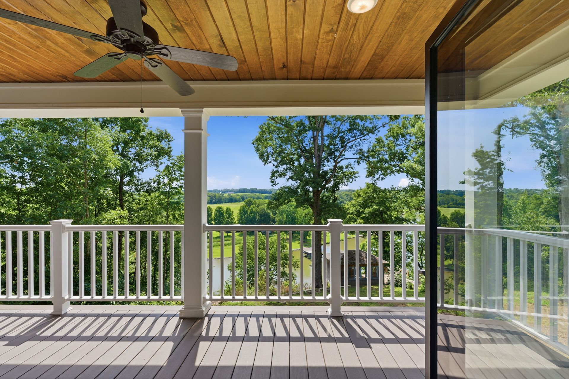 Covered porch with white railing, wood ceiling, and open view of green fields and trees.