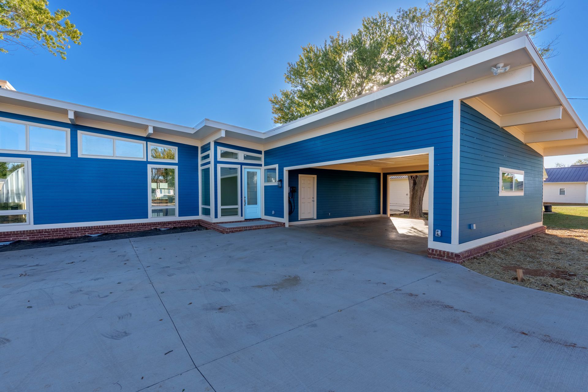 Blue house exterior with carport, wide driveway, and white-trimmed windows under a sunny sky