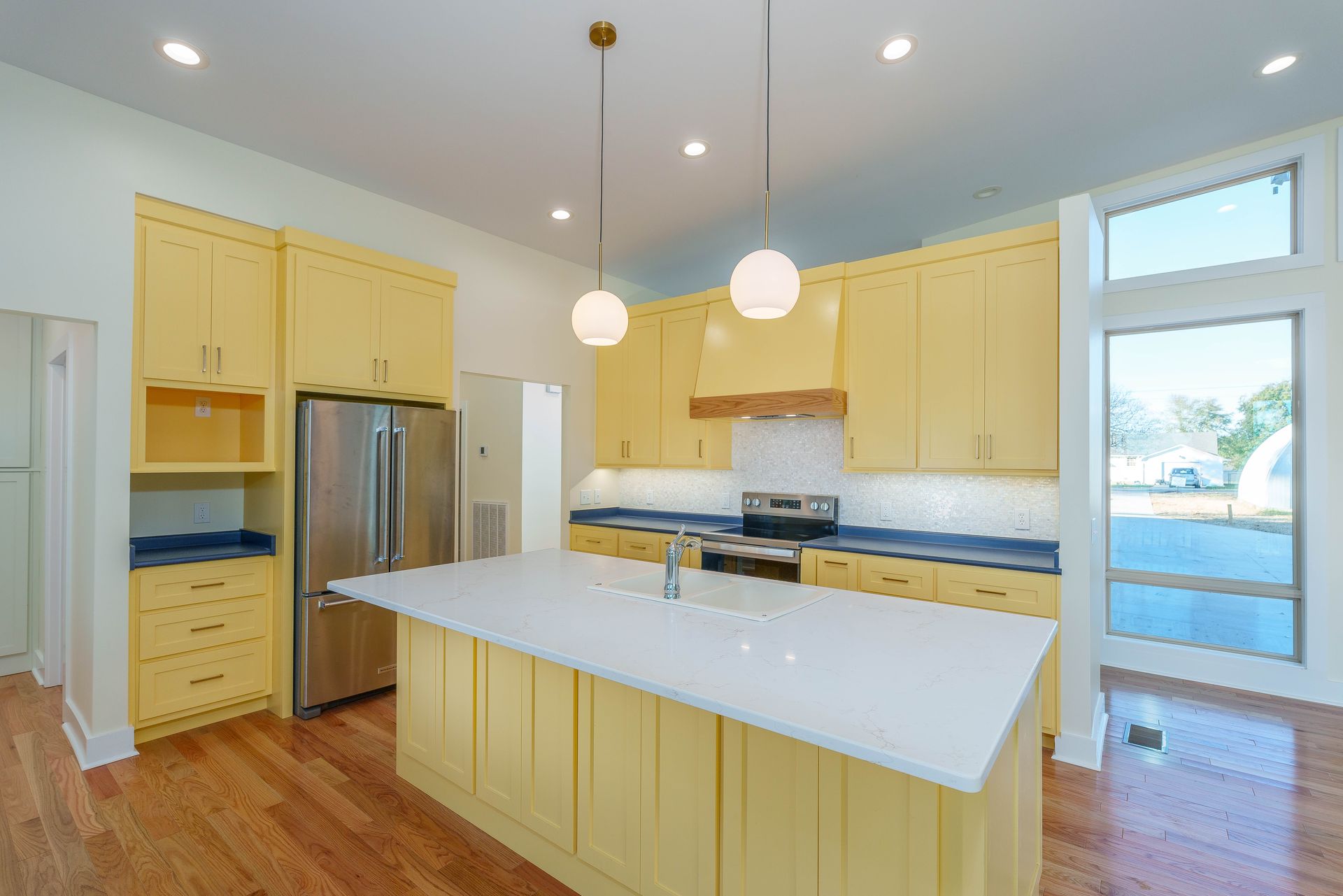 Bright kitchen with yellow cabinets, stainless fridge, white island, and hanging pendant lights.
