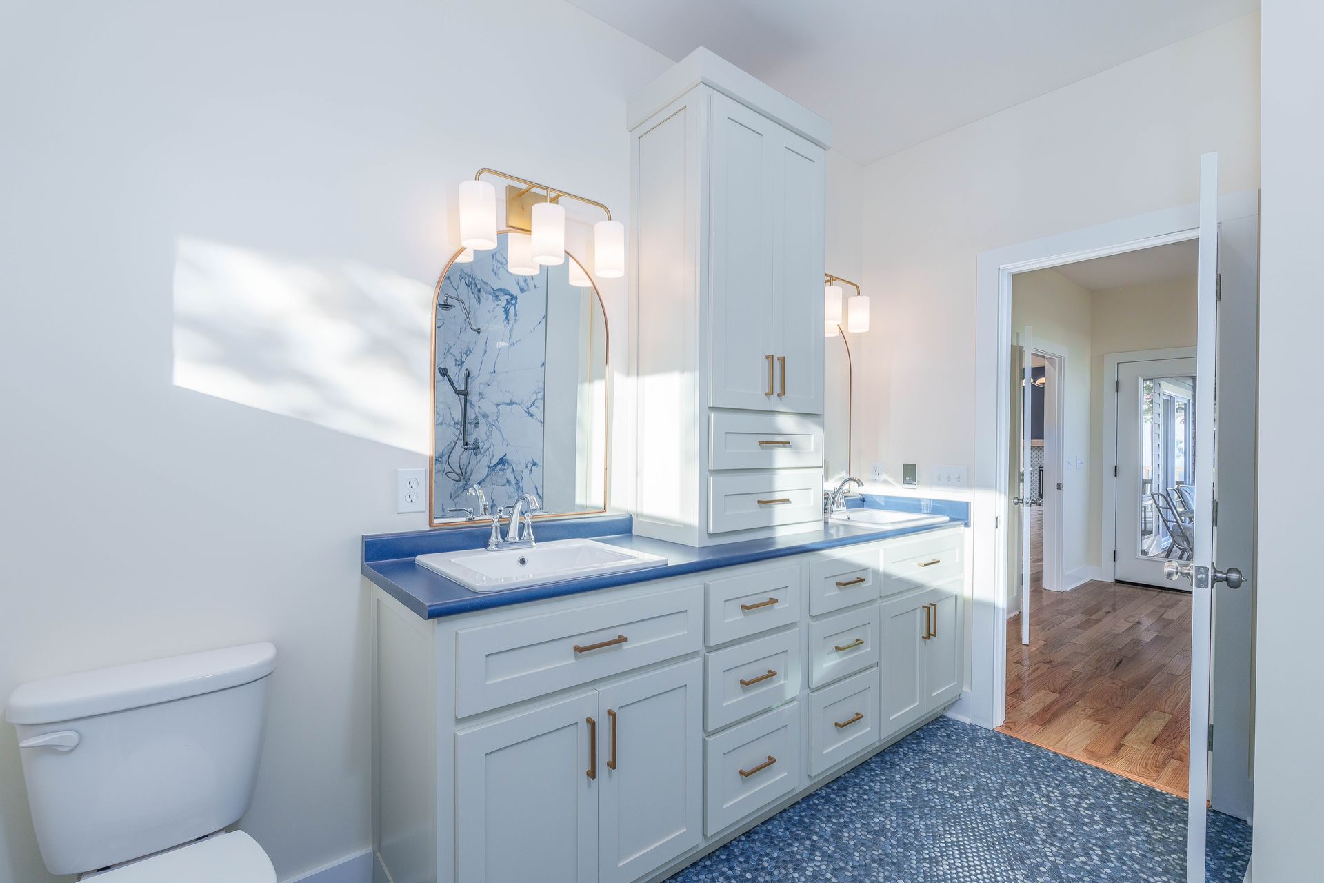 Bright white bathroom with vanity, blue countertop, marble mirror, toilet, and hallway beyond