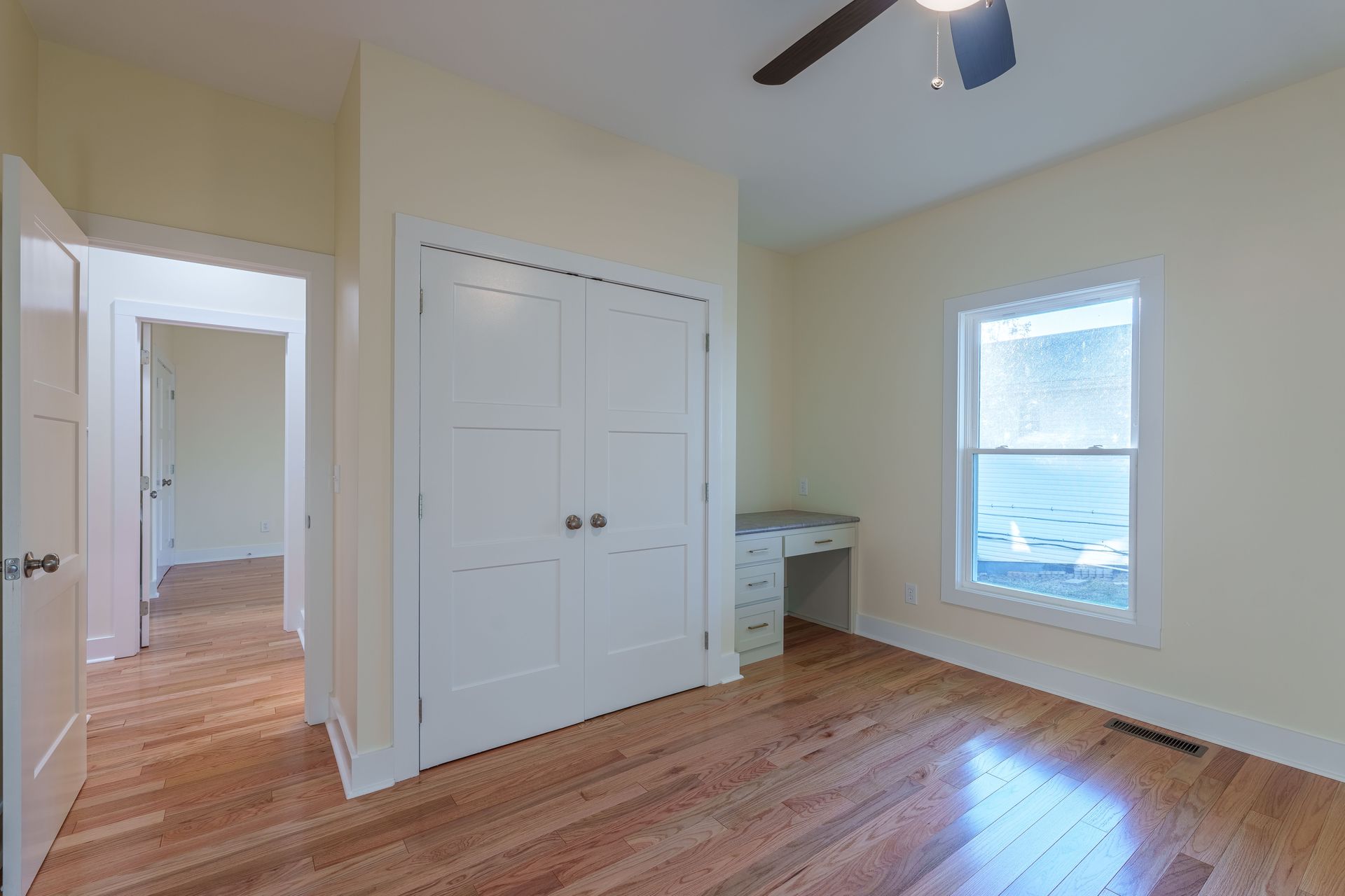 Empty bedroom with hardwood floors, white closet doors, desk, and window with blinds