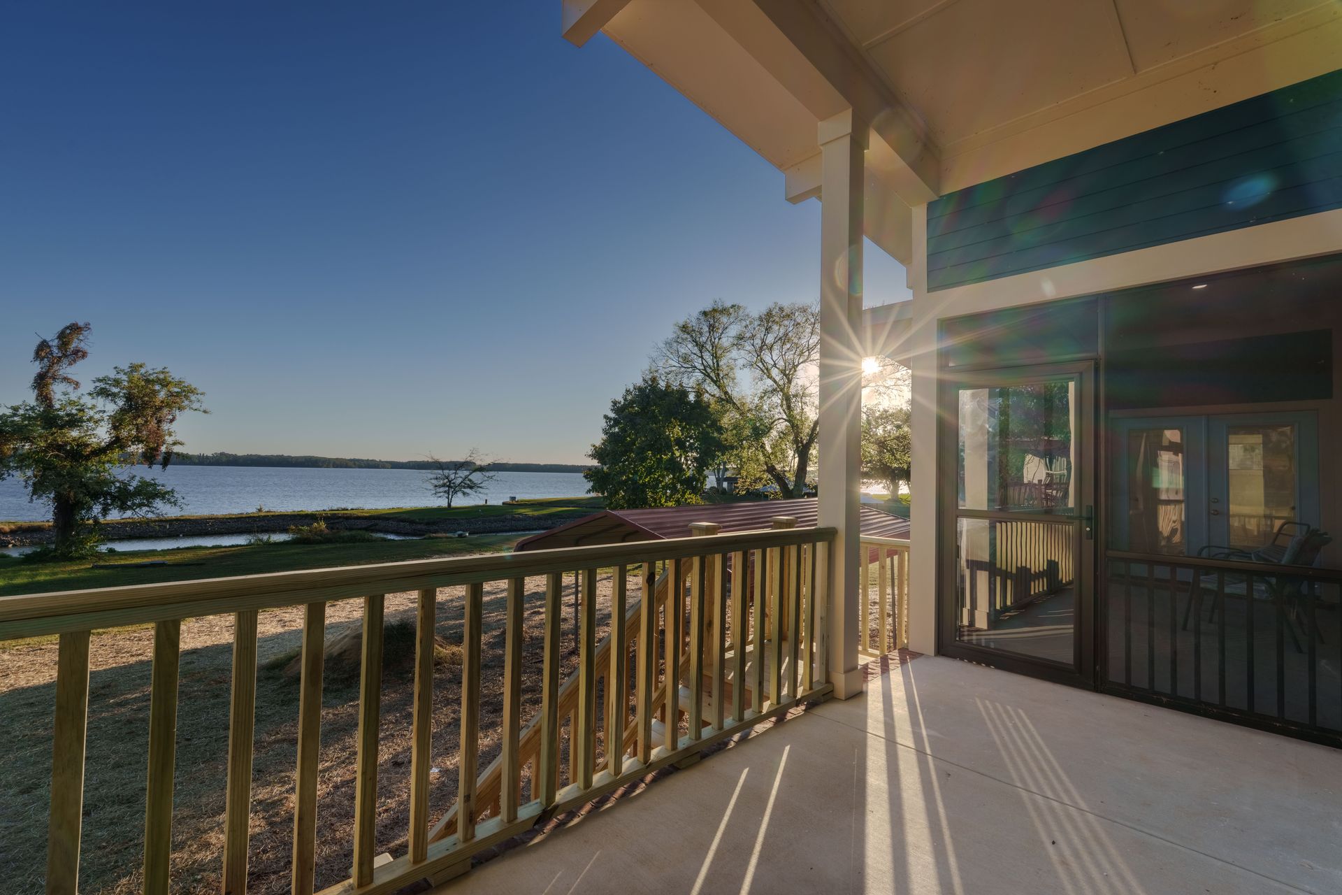 Sunlit porch overlooking a lake, with wooden railing, trees, and sunset reflections on the water.
