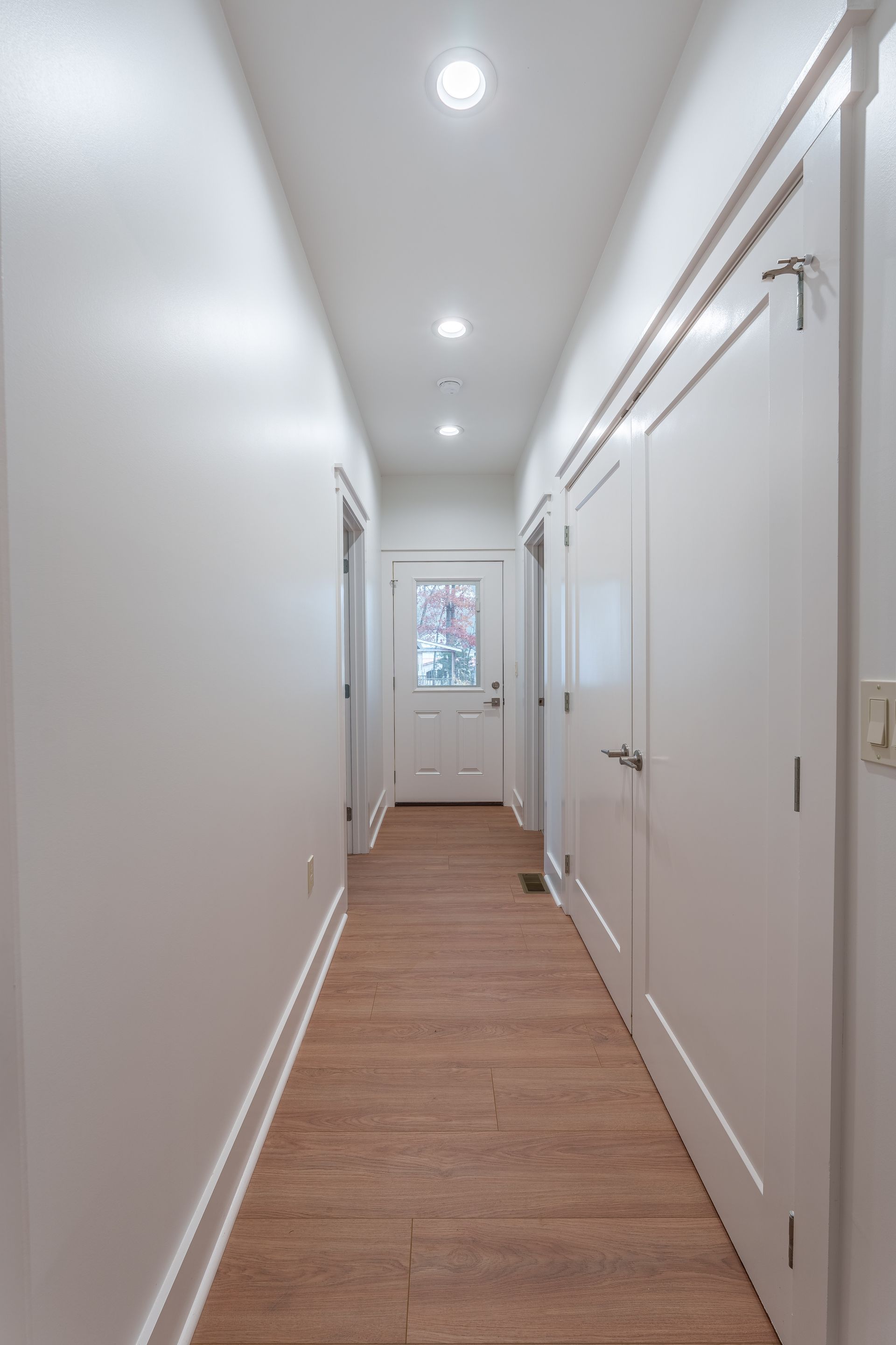 Narrow white hallway with wood floor, recessed lights, and a window at the far end.