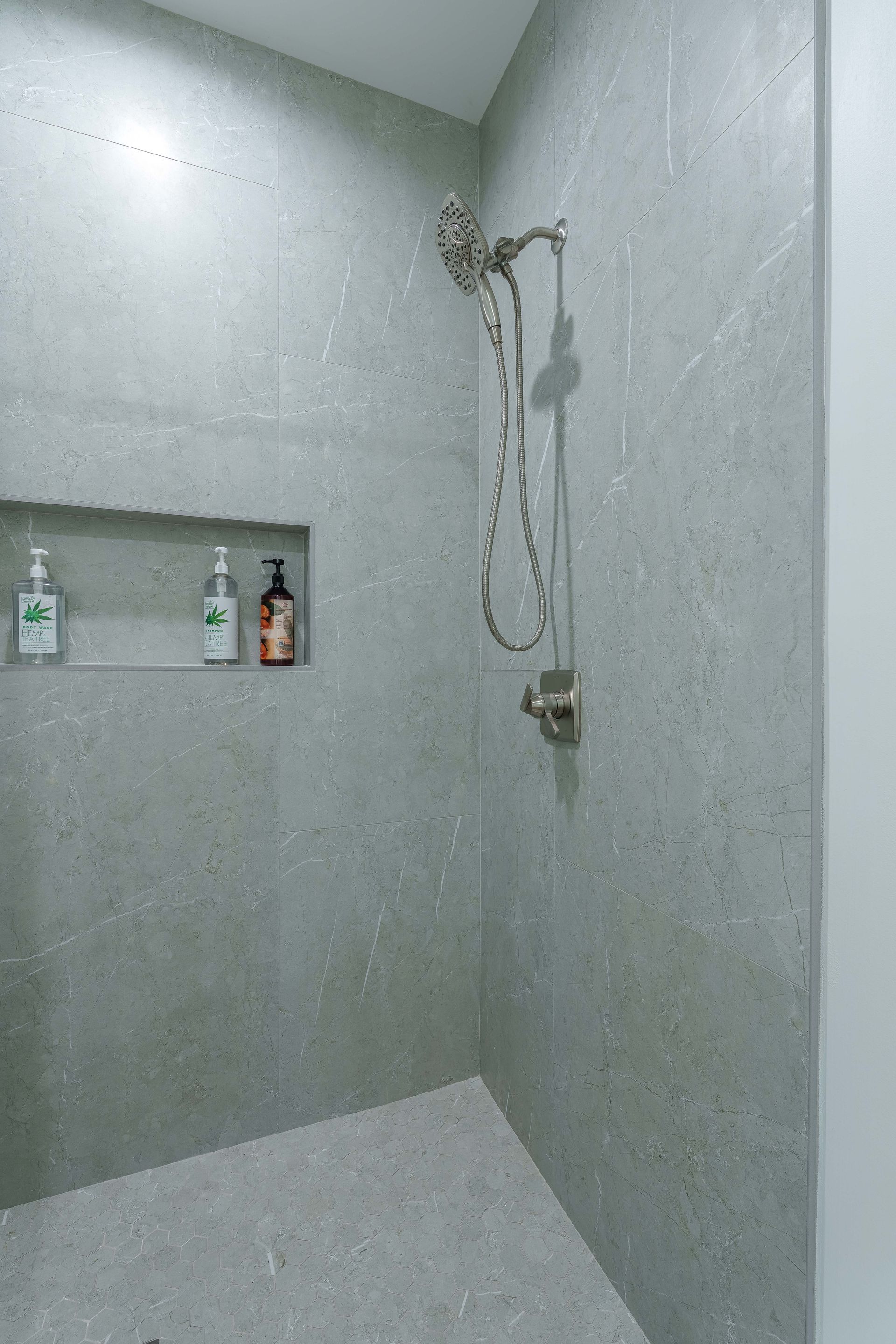 Empty gray shower stall with handheld showerhead, hose, and built-in shelf holding bottles
