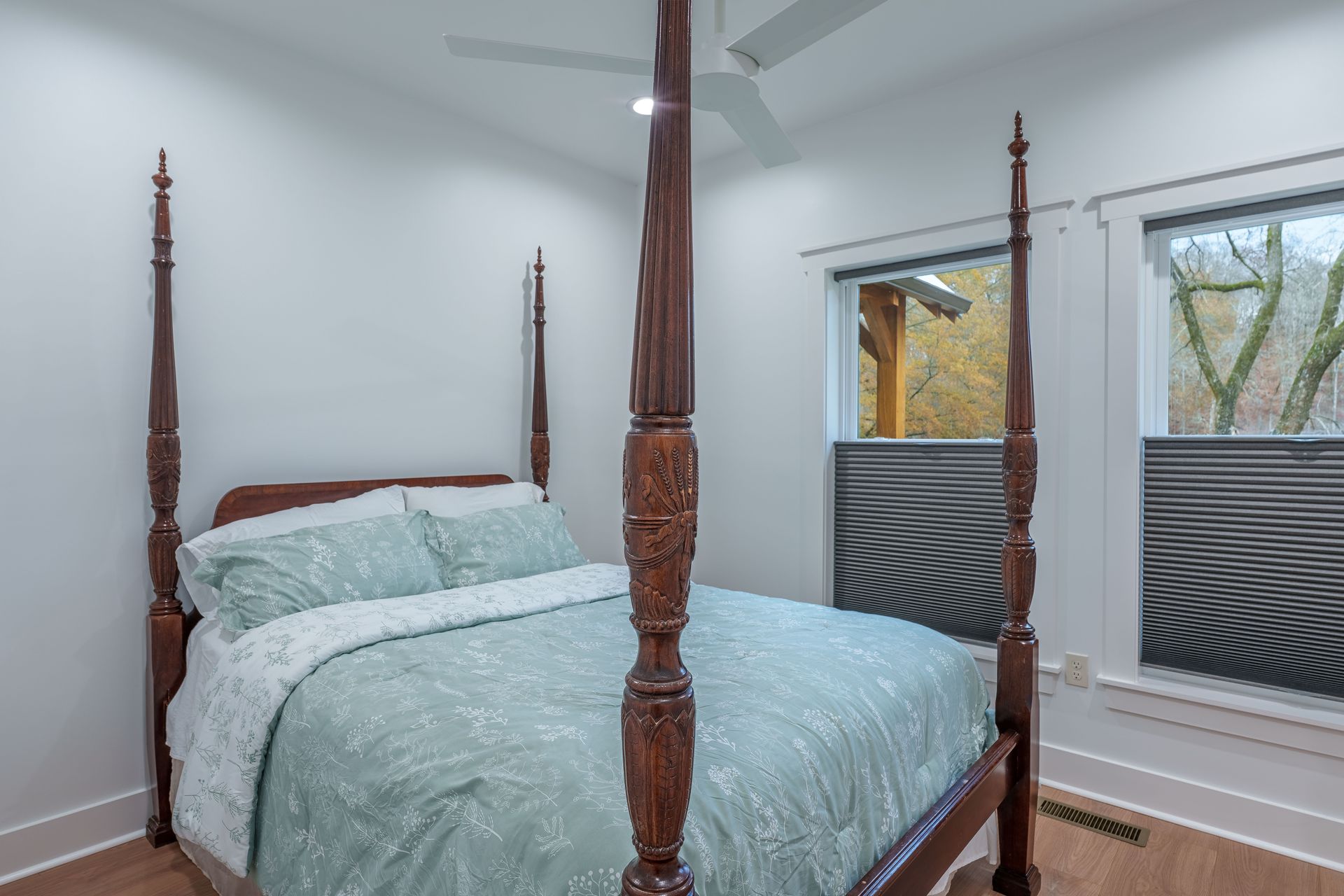 Four-poster bed with pale blue bedding in a bright bedroom, beside large windows with patterned shades