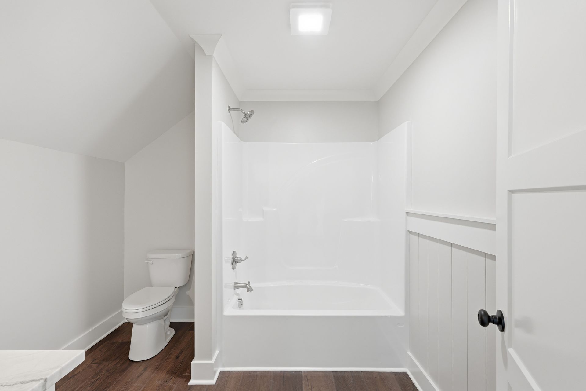 Bright white bathroom with tub-shower, toilet, and wainscoting on wood-look floor.