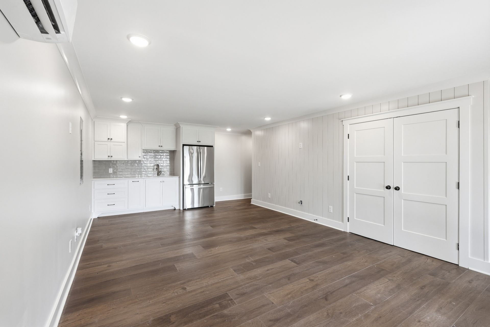 Bright empty open-plan room with white walls, dark wood floors, kitchen, and double doors