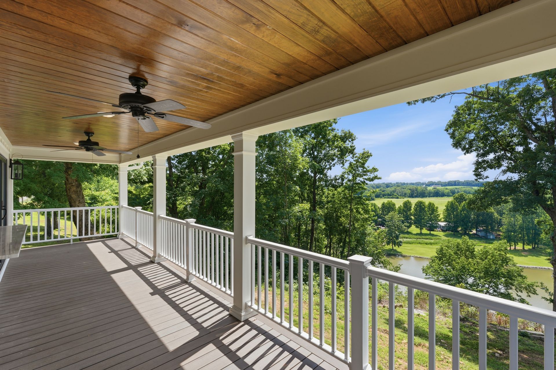 Covered porch overlooking green fields and trees, with white railings and a wood ceiling.