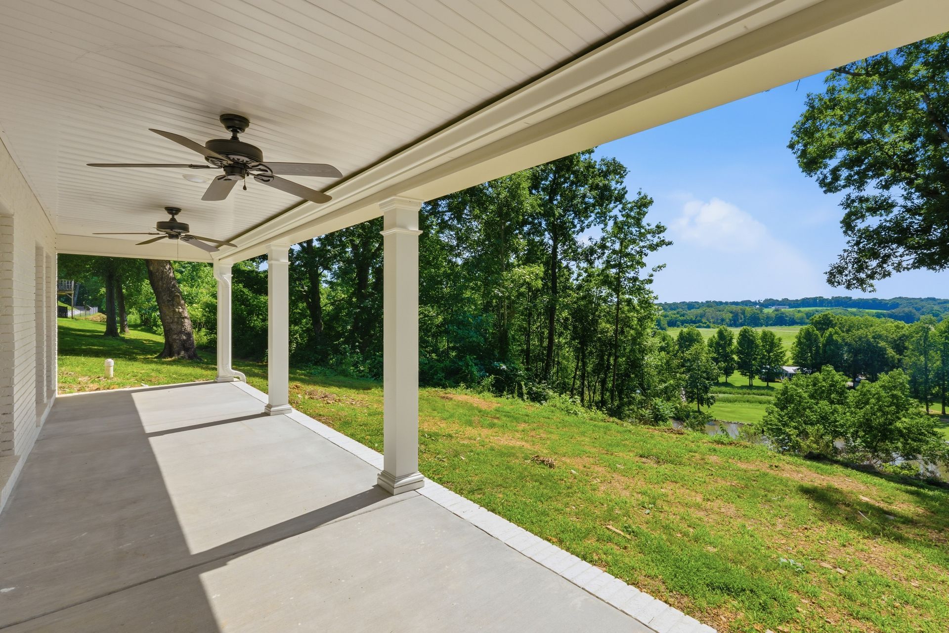 Covered porch with ceiling fan overlooking a grassy yard and wooded landscape on a sunny day