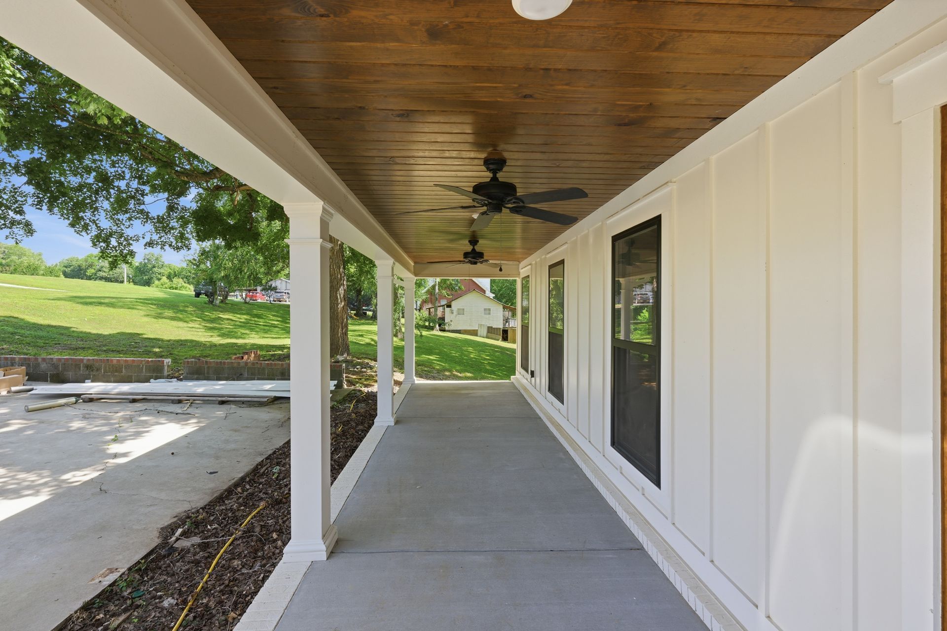 Covered front porch with concrete floor, white columns, wood ceiling, and ceiling fan beside a landscaped yard.