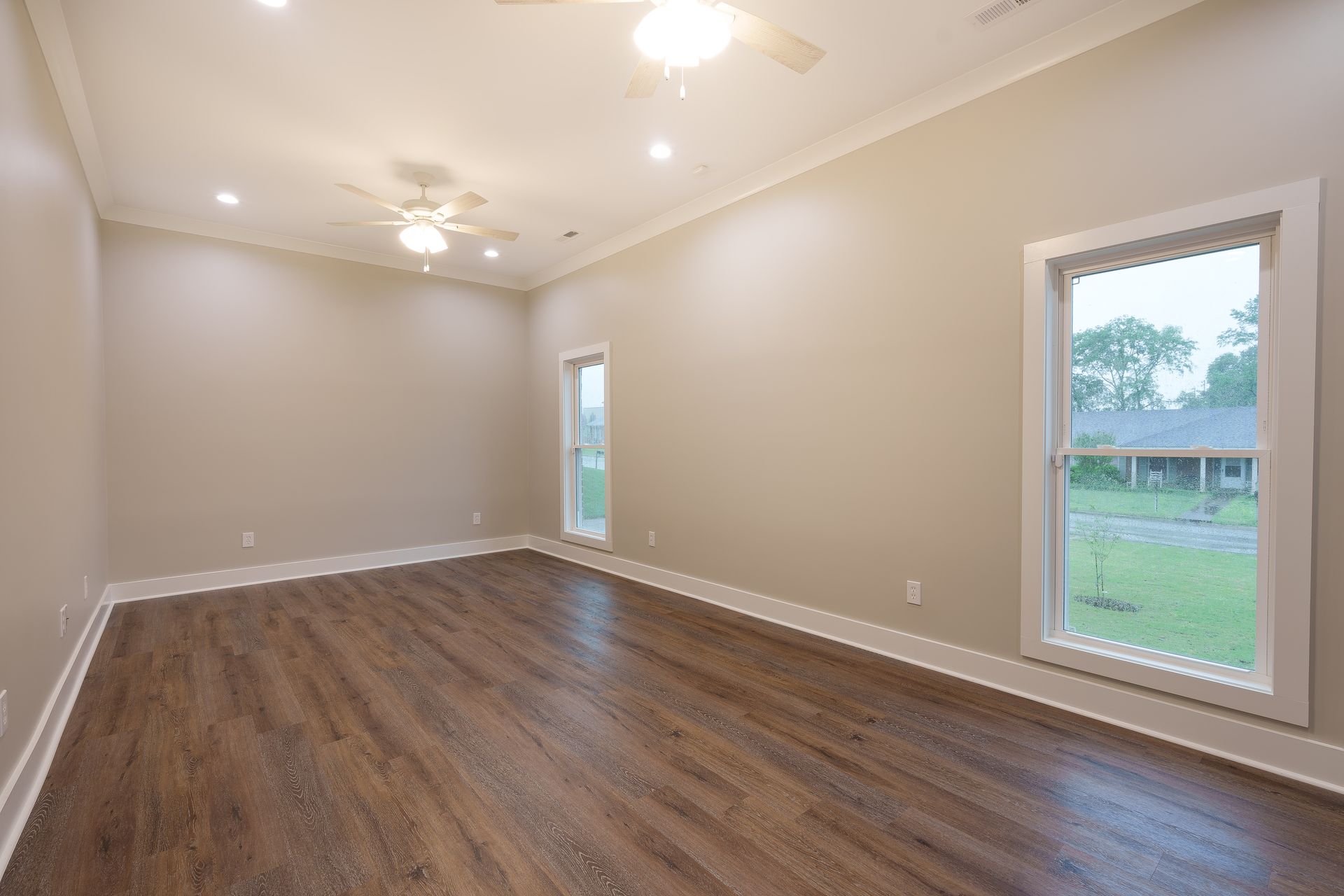 Empty beige living room with wood floors, recessed lights, and two windows overlooking a yard