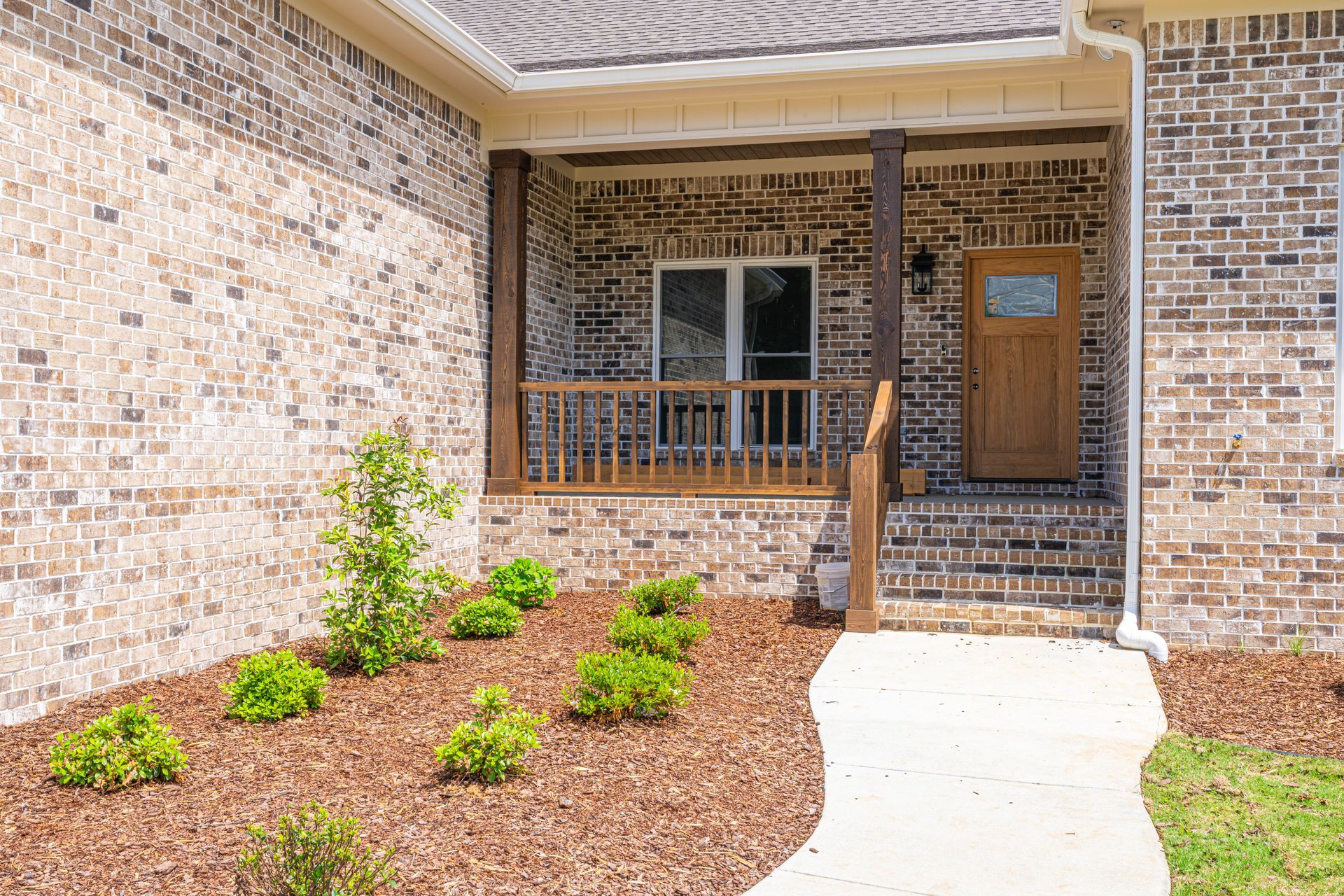 Brick house front porch with window, wooden door, and landscaped mulch bed along a concrete walkway