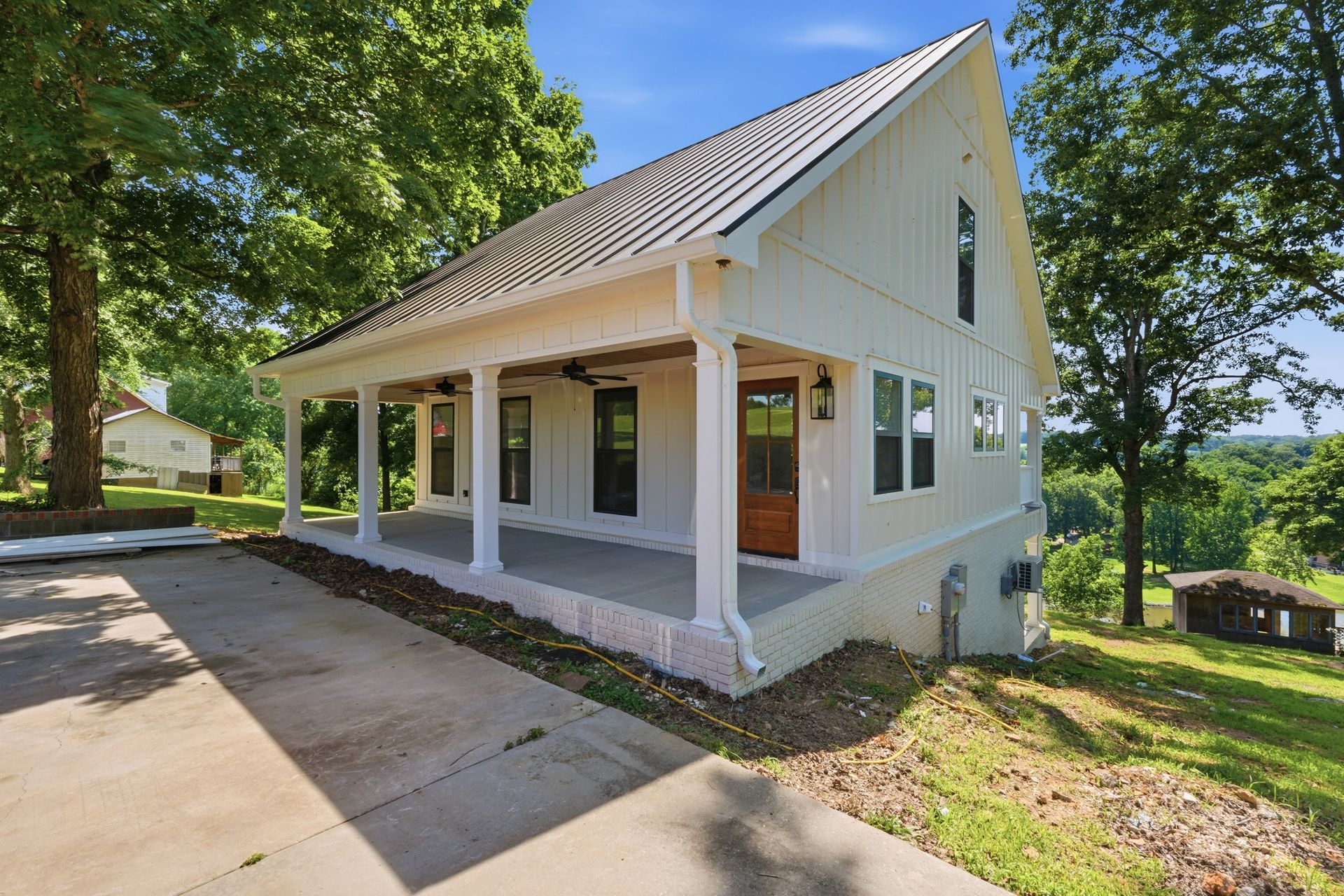 White two-story house with wraparound porch, driveway, and trees in a sunny yard