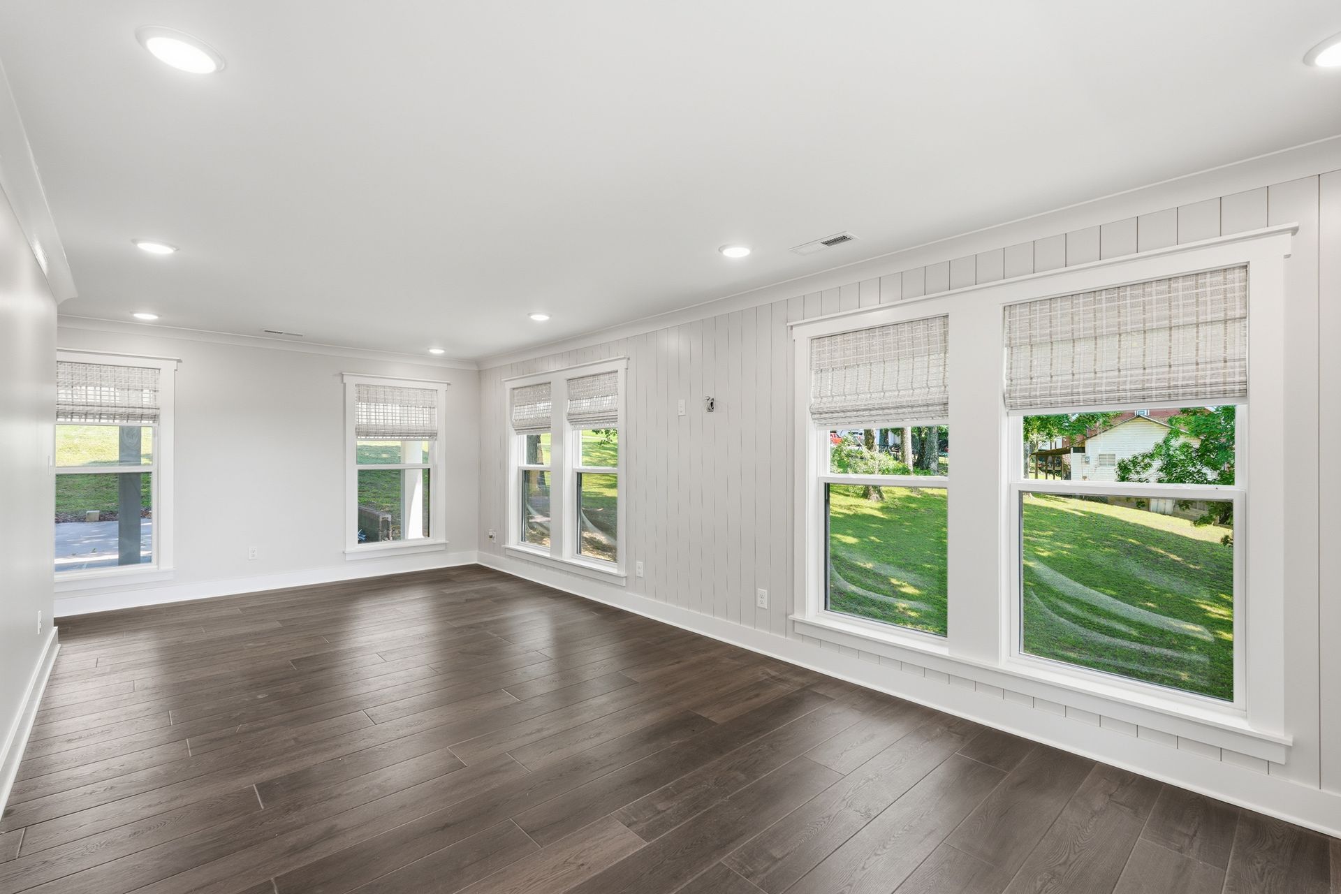 Bright empty room with white walls, dark wood floors, and large windows overlooking a green lawn